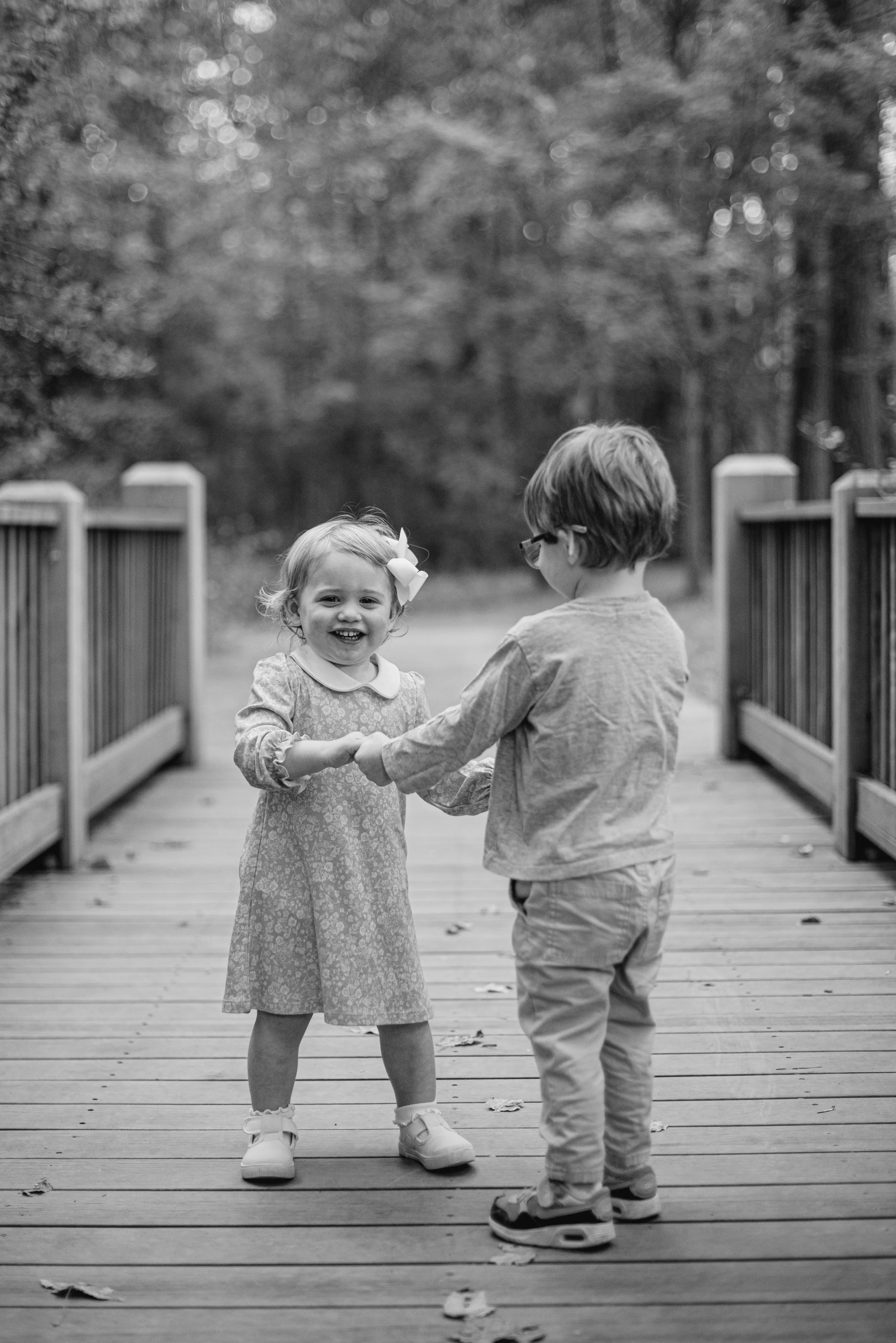 Two young children, a girl and a boy, are standing on a wooden bridge outdoors, holding hands and smiling. The girl wears a dress with a bow in her hair, and the boy is wearing glasses and a long-sleeved shirt. The background features trees and blurr
