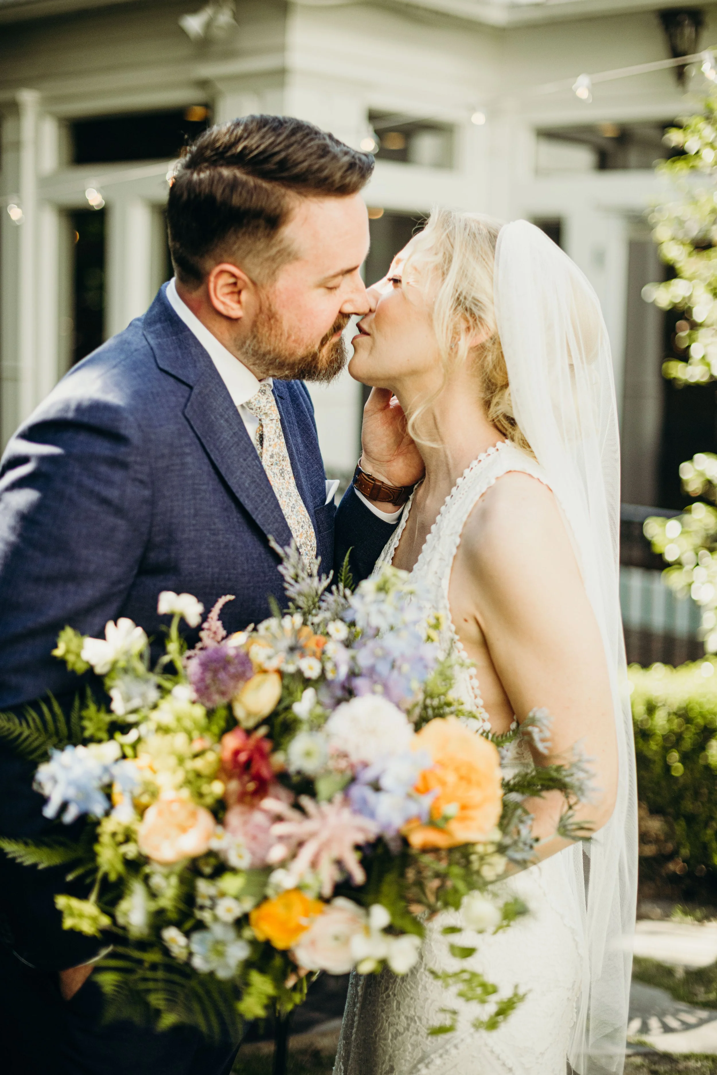 A bride and groom share a kiss outdoors during a wedding ceremony, with a large bouquet of colorful flowers in the foreground.