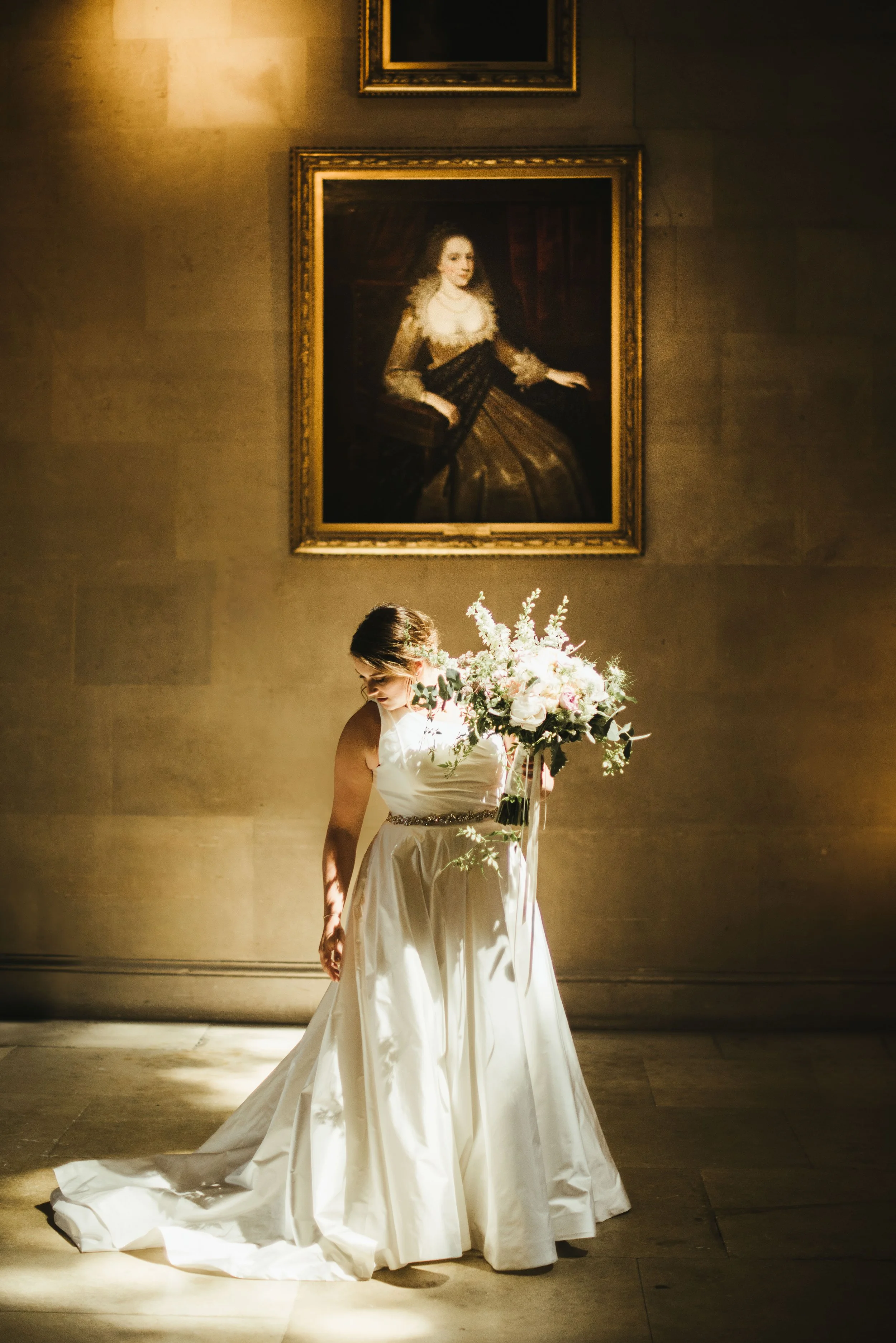 A woman in a white wedding dress holding a bouquet of flowers, standing on a stage with mural lighting. A large, framed portrait painting of a historical woman hangs on the wall behind her.