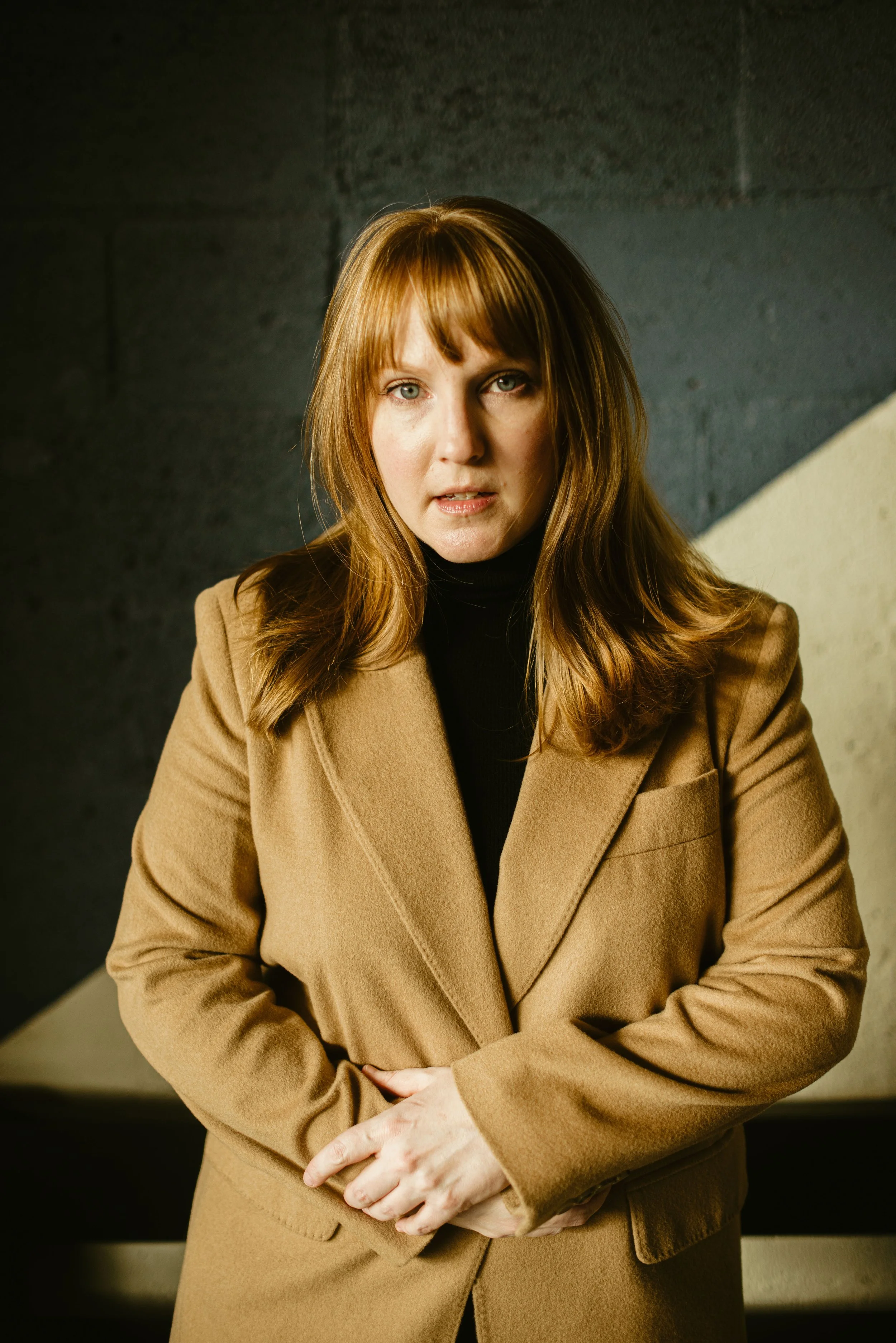 A woman with long, wavy auburn hair wearing a tan coat and black turtleneck, standing indoors against a dark wall with a concrete or brick texture.