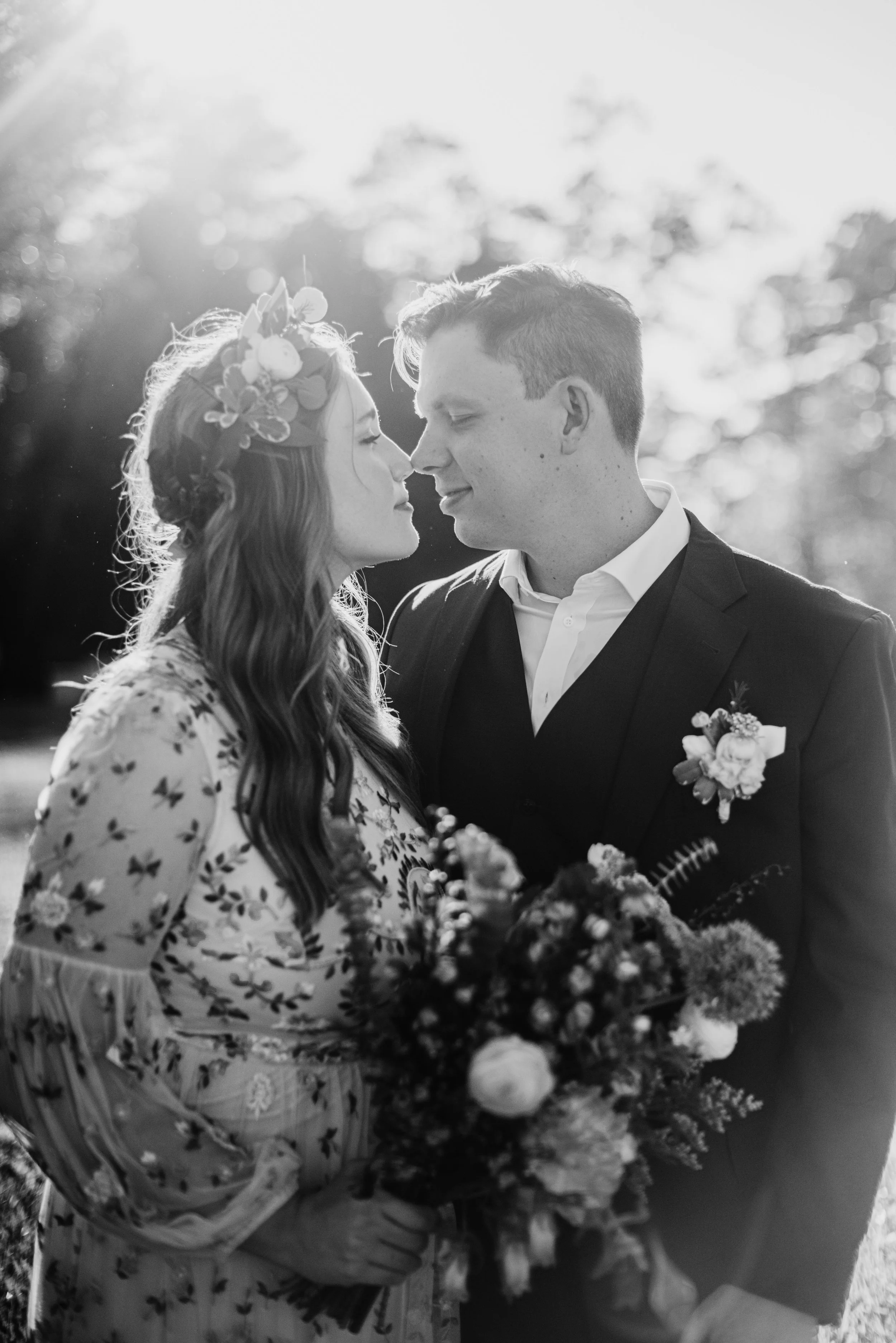 A black and white photo of a couple dressed for a wedding, the woman wearing a floral crown and holding a bouquet, and they are touching foreheads with eyes closed, outdoors with sunlight filtering through trees.