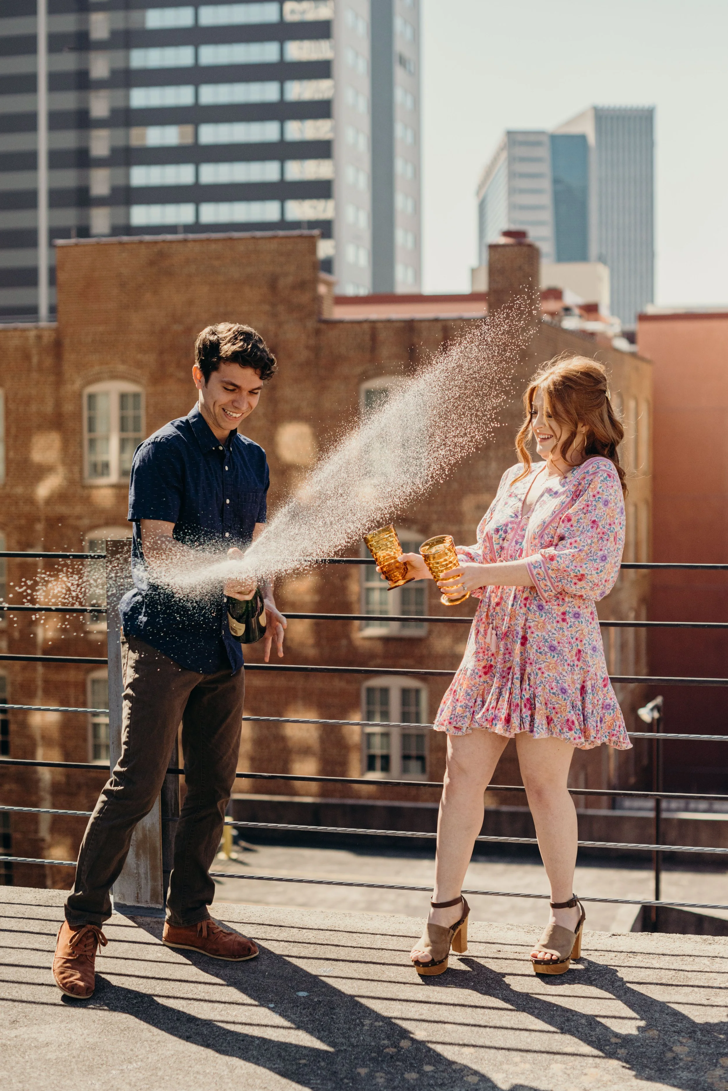A young man in a blue shirt and brown pants laughing and spraying champagne into a woman's face on a rooftop with city buildings in the background. The woman is in a floral dress and high heels, smiling and holding two glasses.