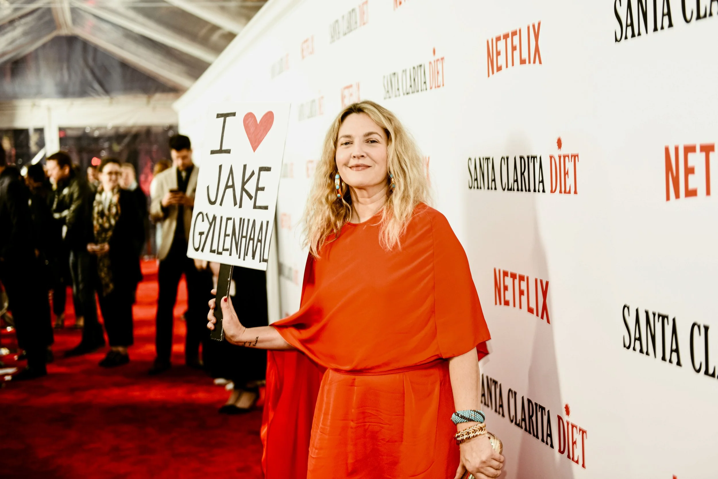 Drew Barrymore wearing a red dress, stands at a red carpet event holding a sign. She is smiling and standing against a white wall with text and logos related to Netflix and Santa Clarita Diet. There are other people in the background.
