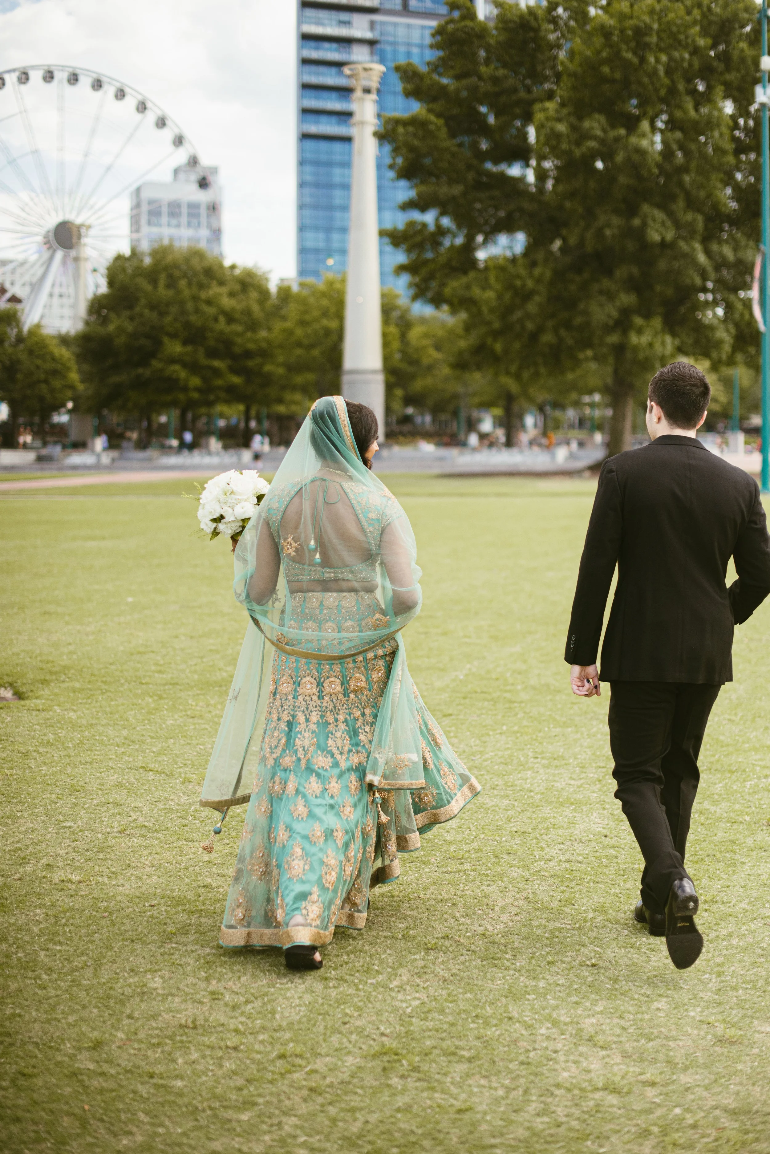 A couple walking outdoors in a park, the woman dressed in traditional South Asian attire holding a bouquet of flowers, and the man in a black suit. In the background are trees, modern buildings, and a Ferris wheel.