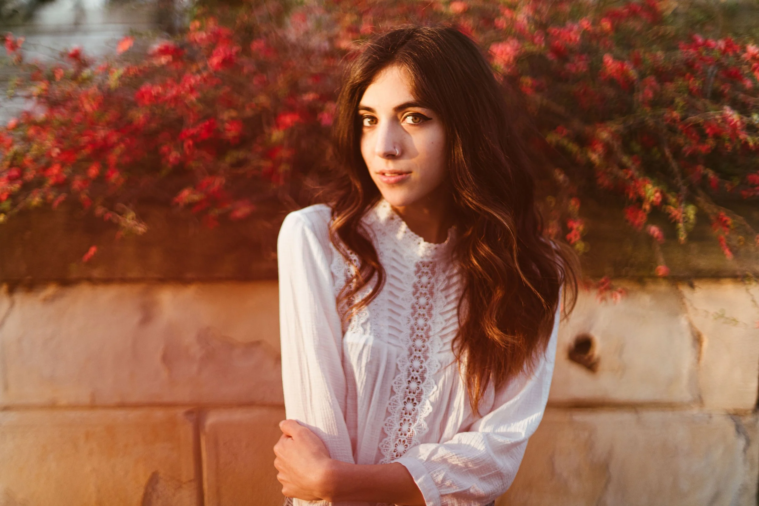 Young woman with long brown hair and light makeup, wearing a white lace blouse, standing outdoors in front of a wall with pink flowers. The photo is taken during golden hour, giving a warm glow.