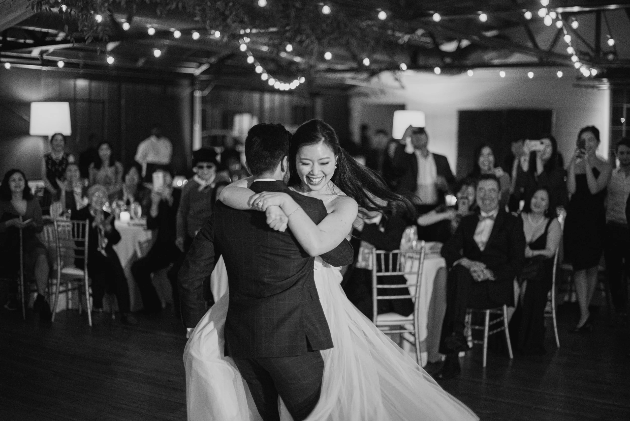 A couple dances closely at a wedding reception with guests watching and taking photos in the background. The reception is decorated with string lights and warm lighting.