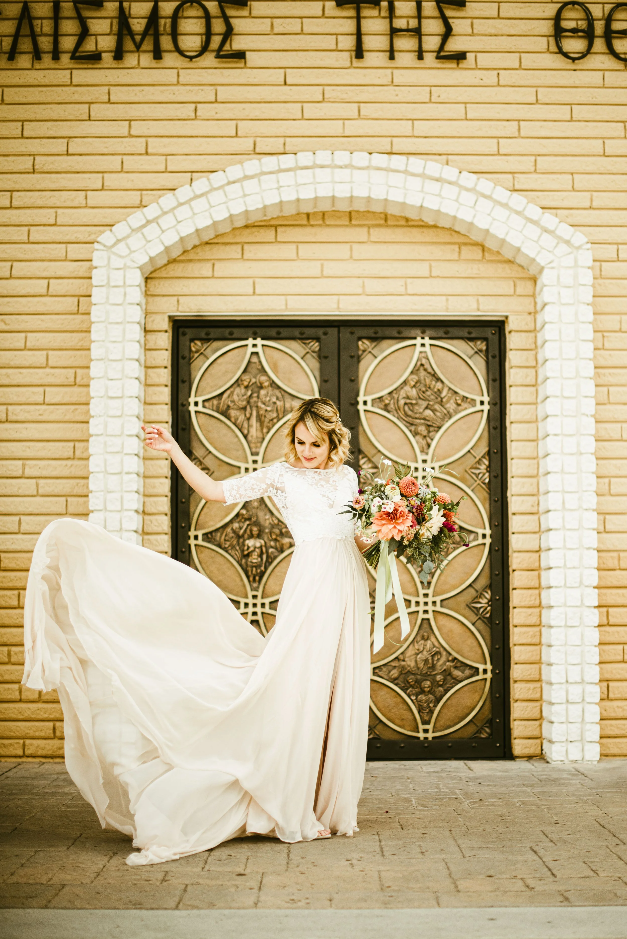 A bride in a white wedding dress holding a bouquet of flowers, standing in front of an ornate metal door with biblical reliefs, across a brick wall with a white arched frame in the background.