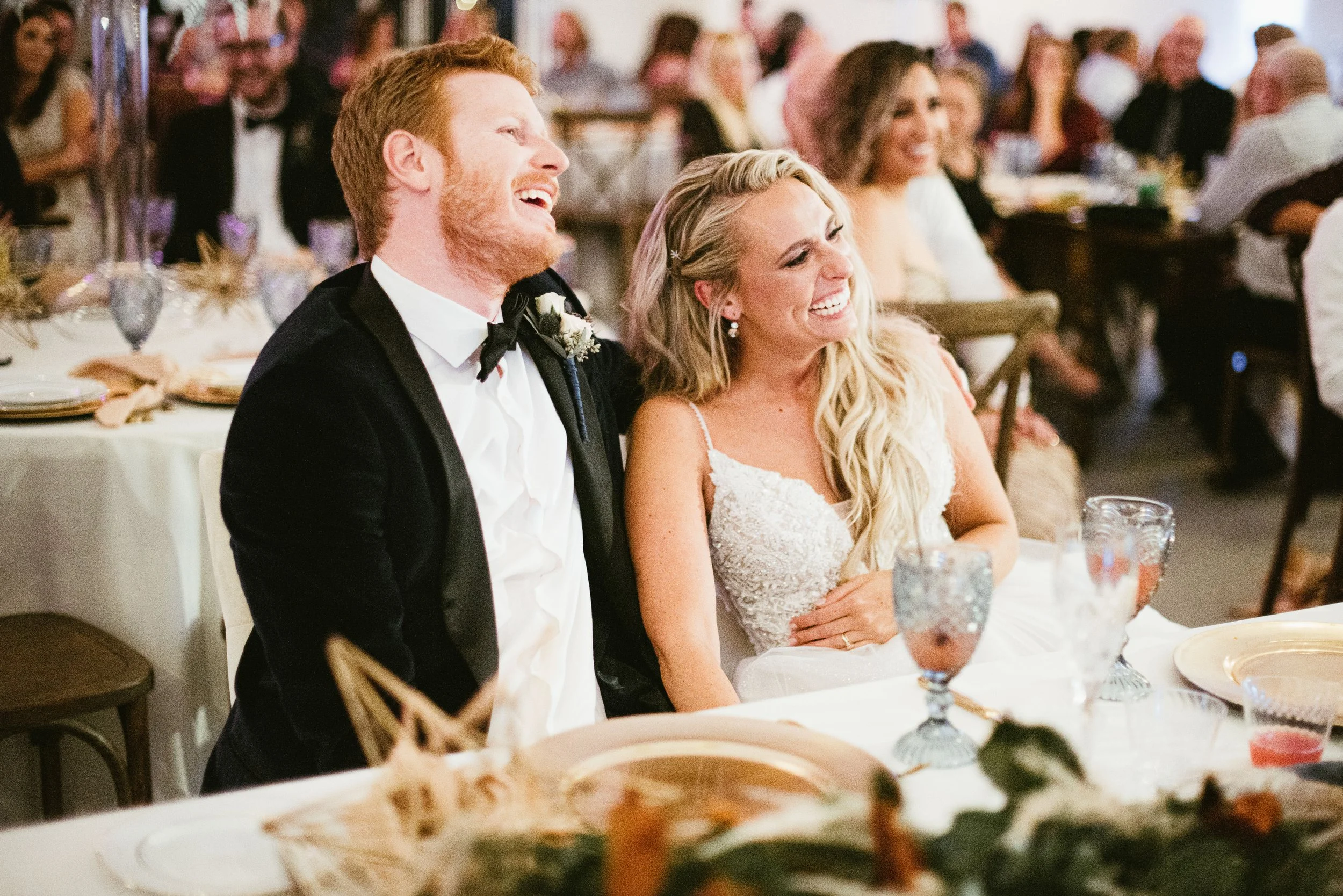 A group of people at a wedding reception, including a man with red hair and a beard wearing a tuxedo, and a woman with long blonde hair in a wedding dress, both smiling and laughing. The background shows other guests seated at tables, and the scene a