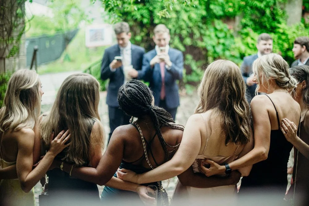 Group of women embracing and socializing outdoors at a gathering, with two men in suits taking photos in the background.