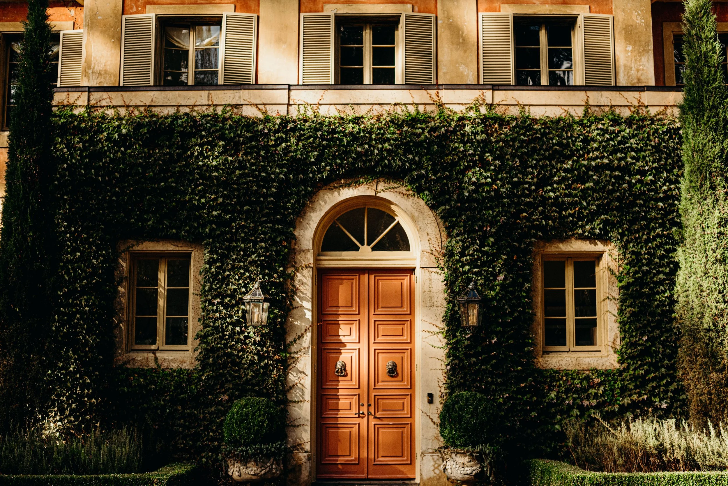 A building's front façade with a wooden double door, surrounded by ivy and greenery, with four tall windows with shutters above and two lanterns flanking the door.