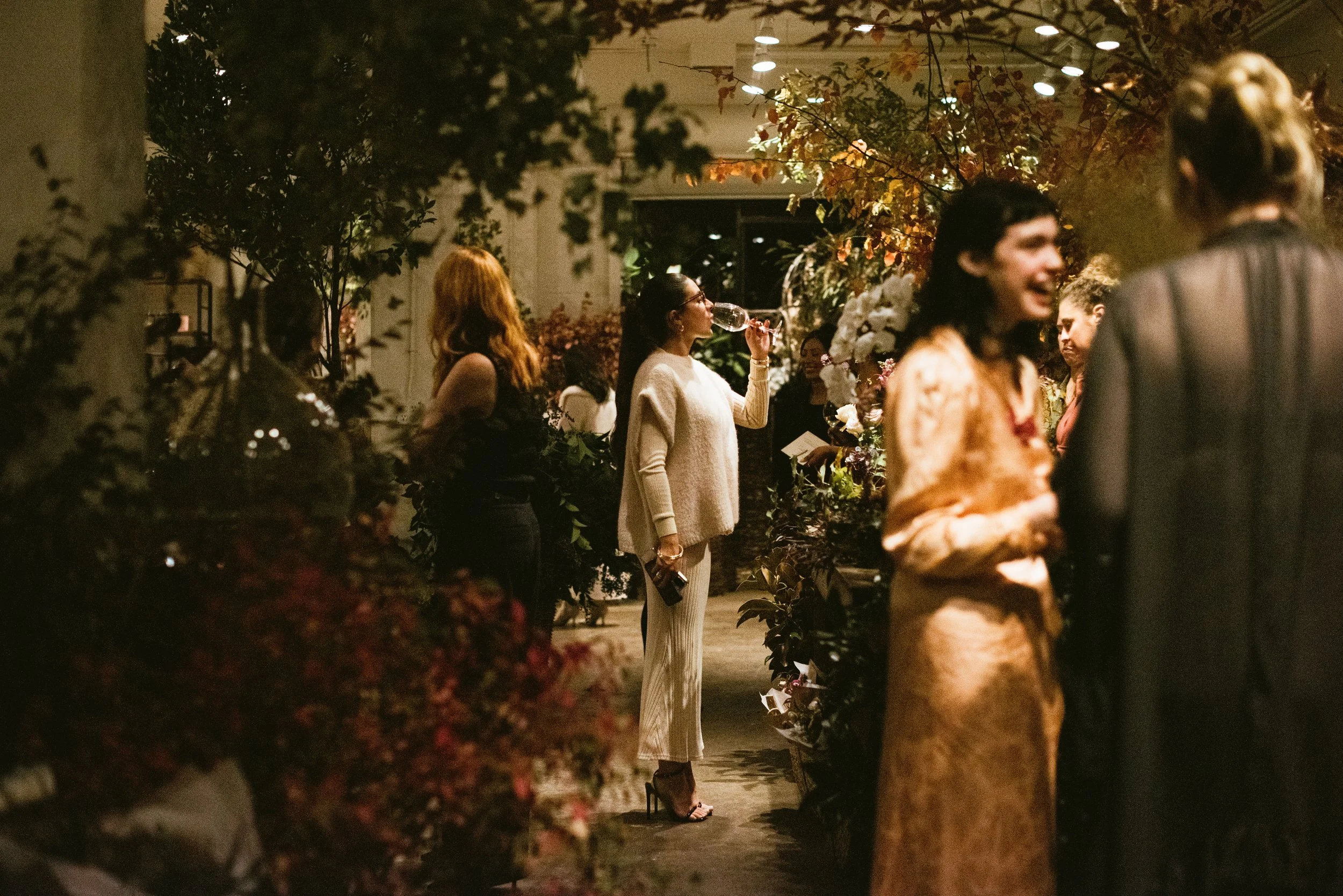 People socializing at an indoor event with plants and warm lighting, including women in stylish outfits, one drinking wine.