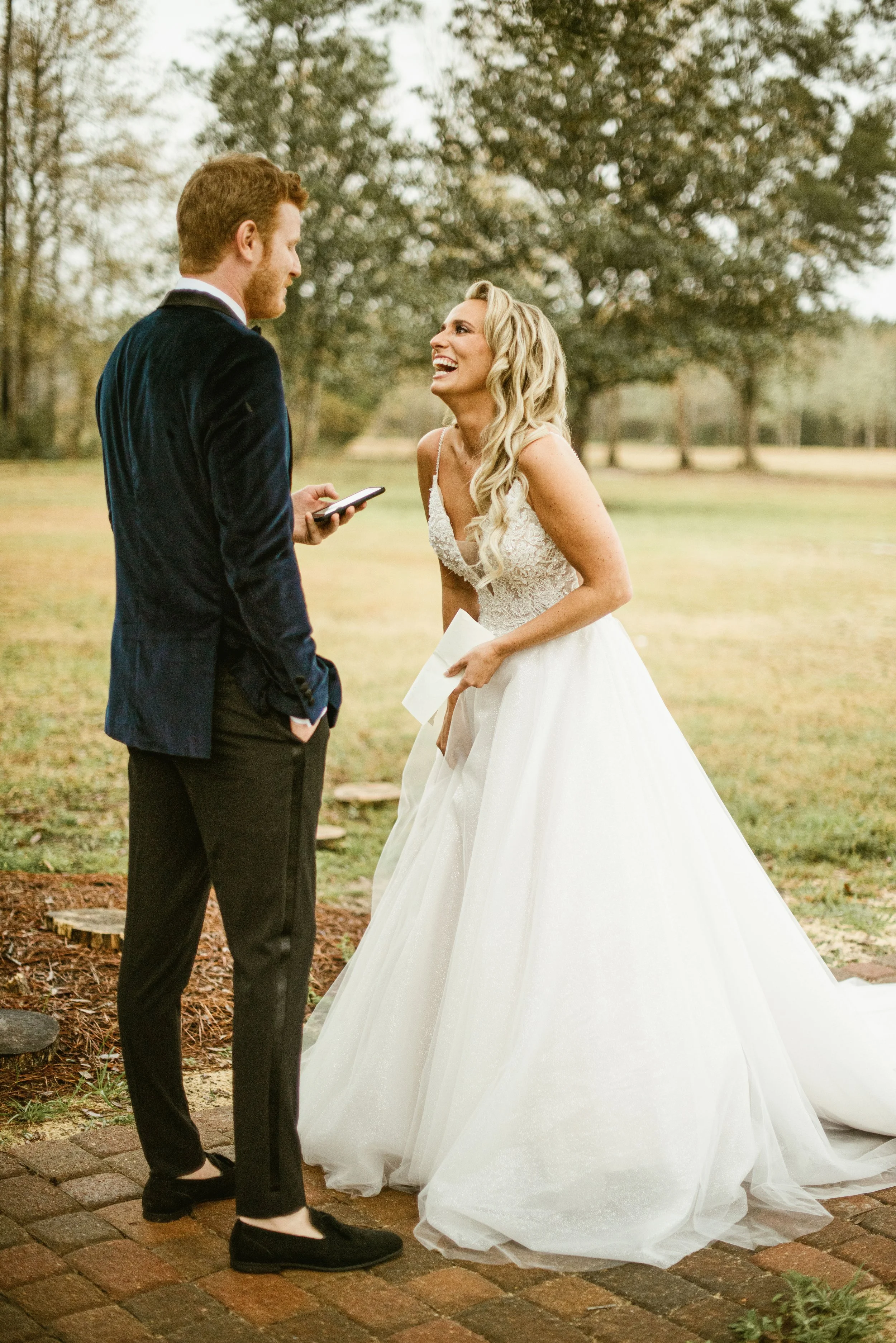 A bride and groom standing outdoors in a park, smiling at each other during their wedding ceremony.