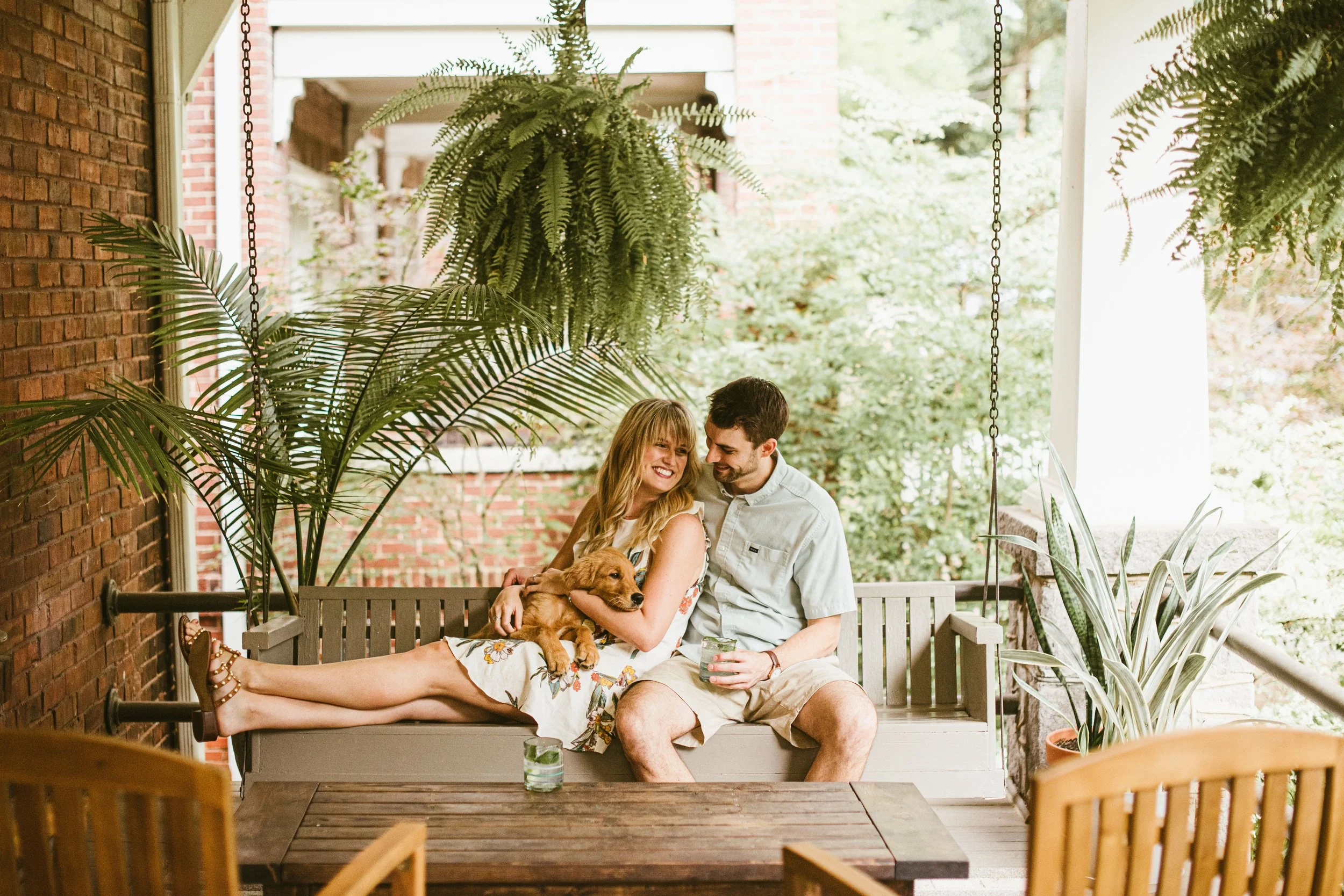 A happy couple sits on a porch swing, holding and cuddling a small brown puppy, surrounded by lush green plants, with a brick wall and trees in the background.