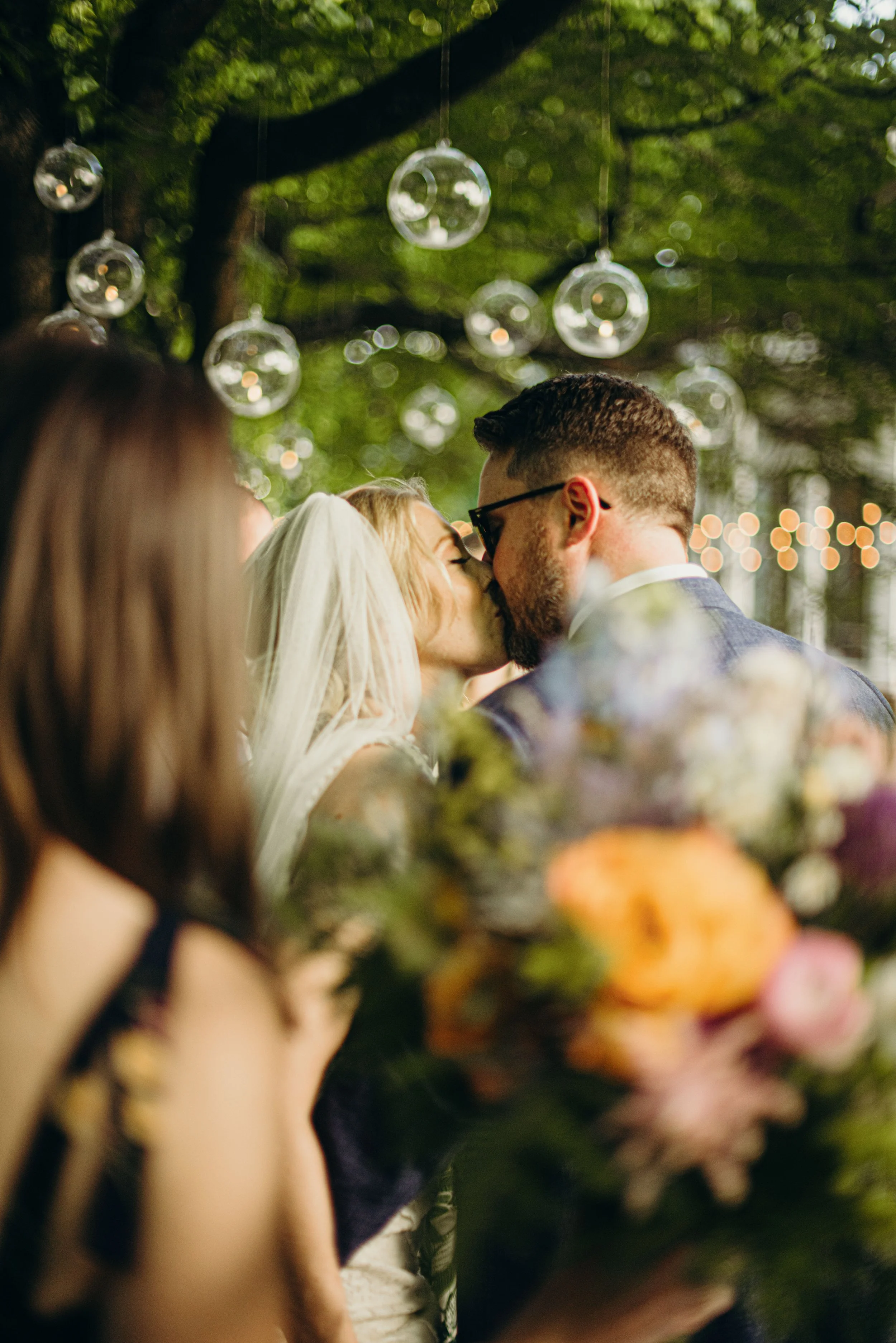 A couple sharing a kiss at an outdoor wedding or celebration surrounded by string lights and hanging glass ornaments, with blurred flowers in the foreground.