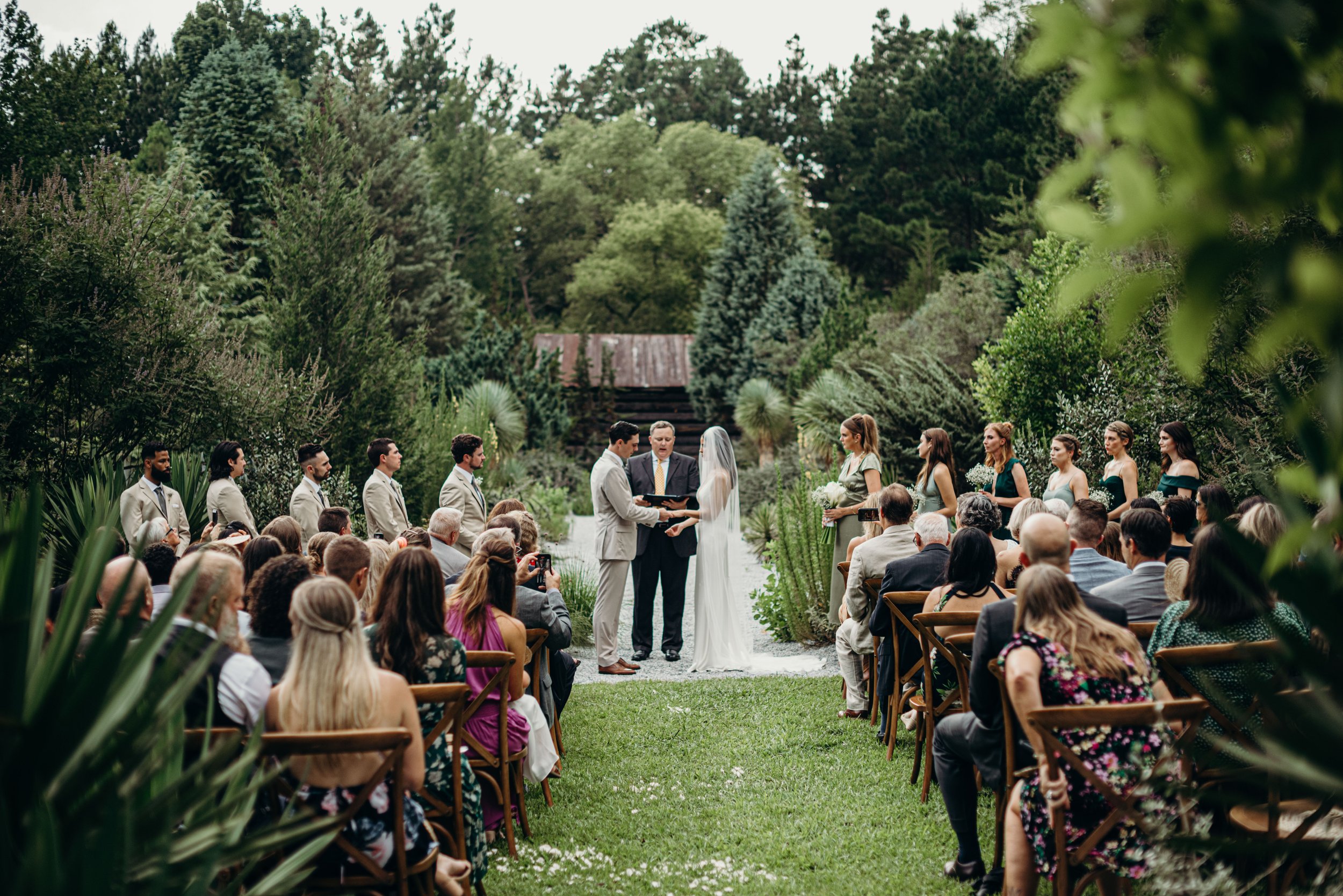 A wedding ceremony taking place outdoors in a lush, green garden with guests seated on either side of the aisle, and the bride and groom standing before an officiant.