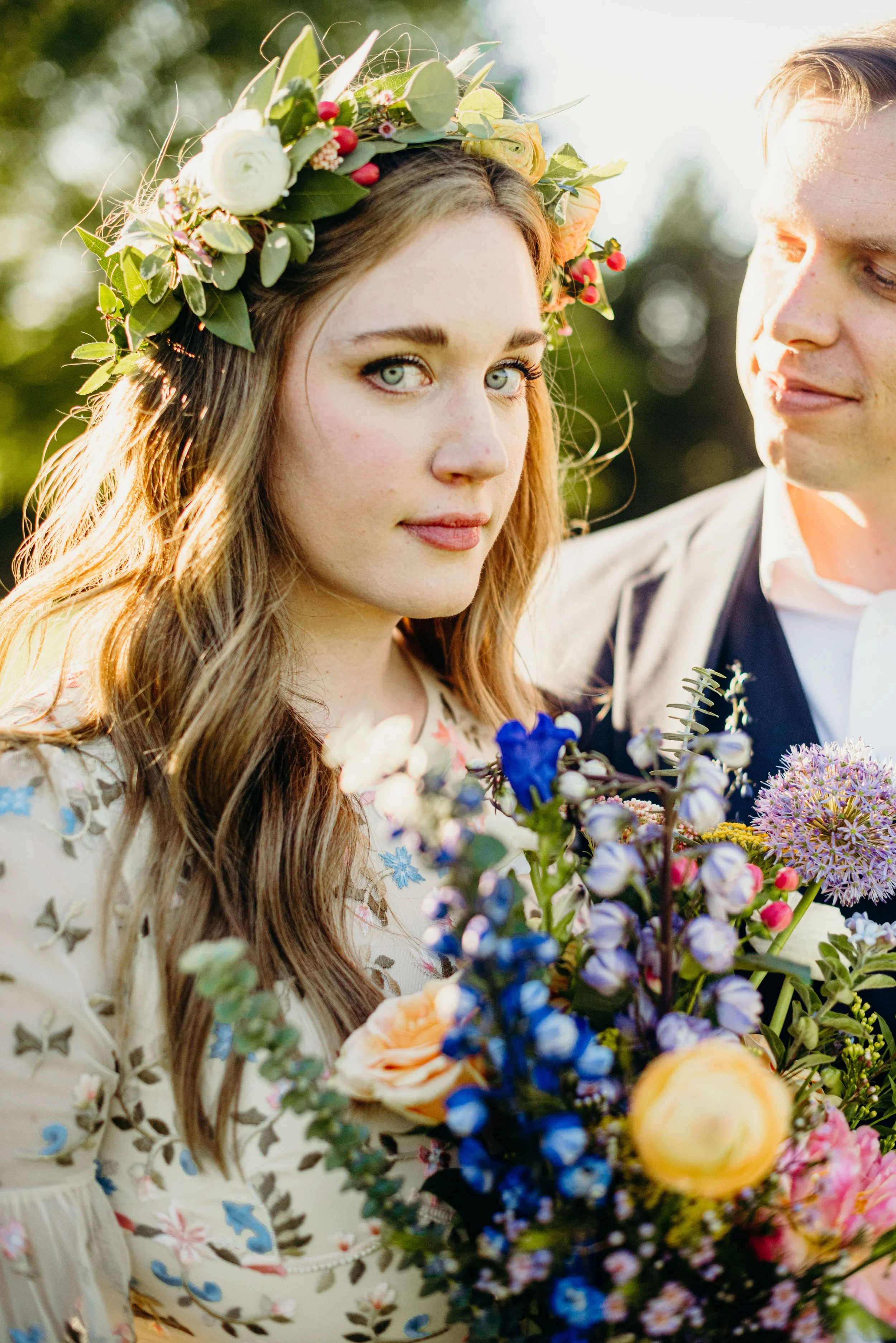 A young woman with blue eyes and long brown hair wearing a floral crown and holding a colorful bouquet of flowers, standing beside a man outdoors during daylight.