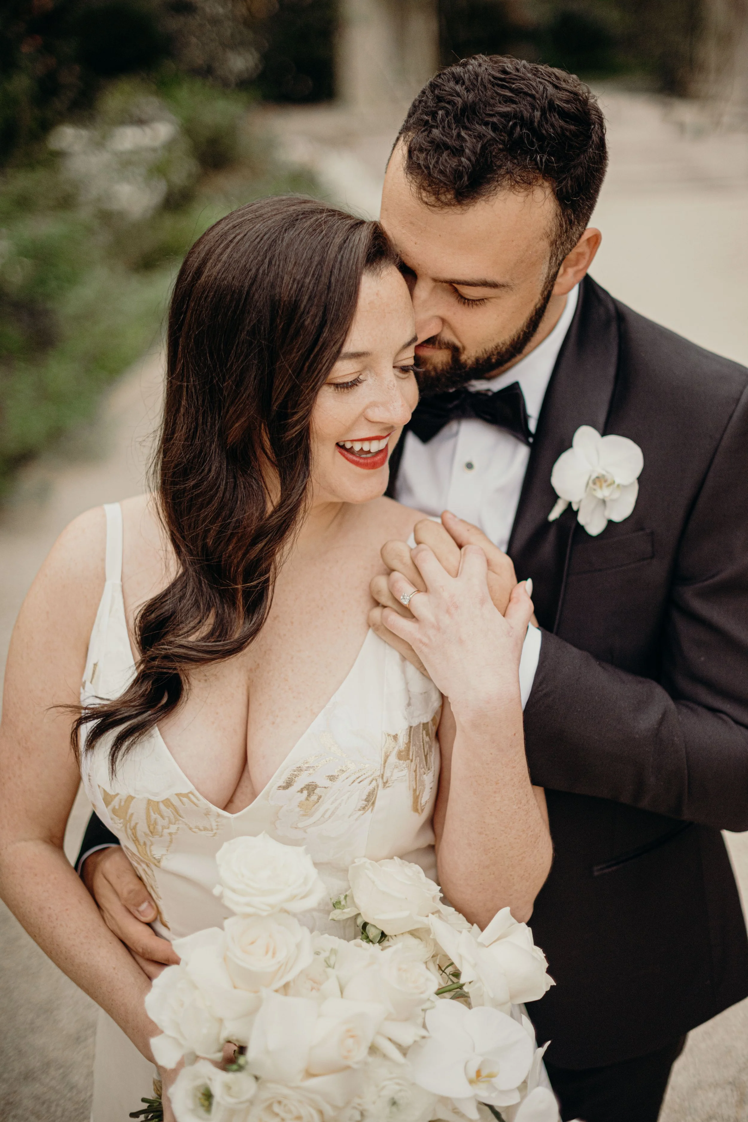 A newlywed couple in wedding attire, smiling and embracing each other outdoors surrounded by greenery, with the bride holding a large bouquet of white and cream roses.