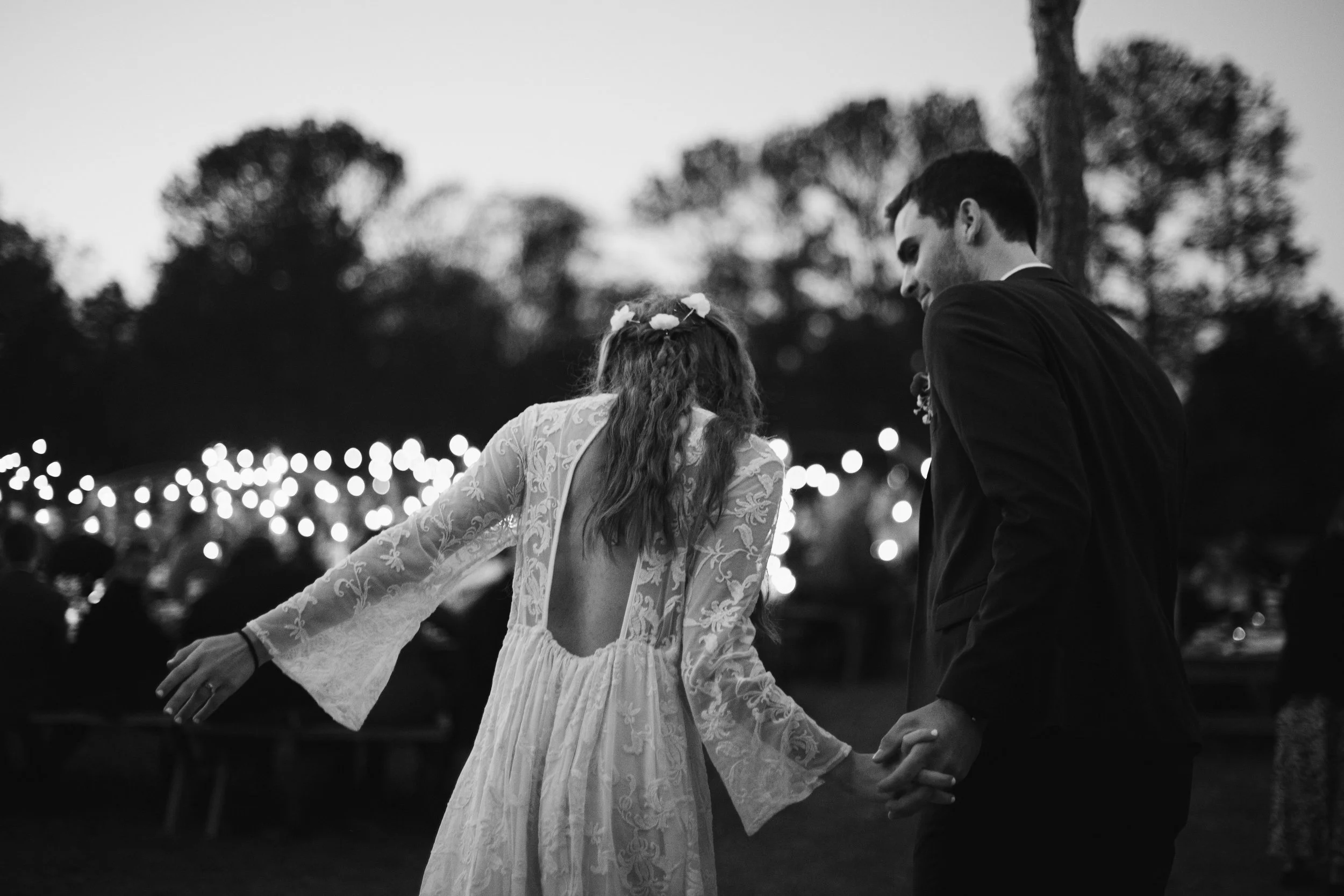 A couple dancing outdoors at twilight, with blurred string lights and trees in the background, black and white photograph.