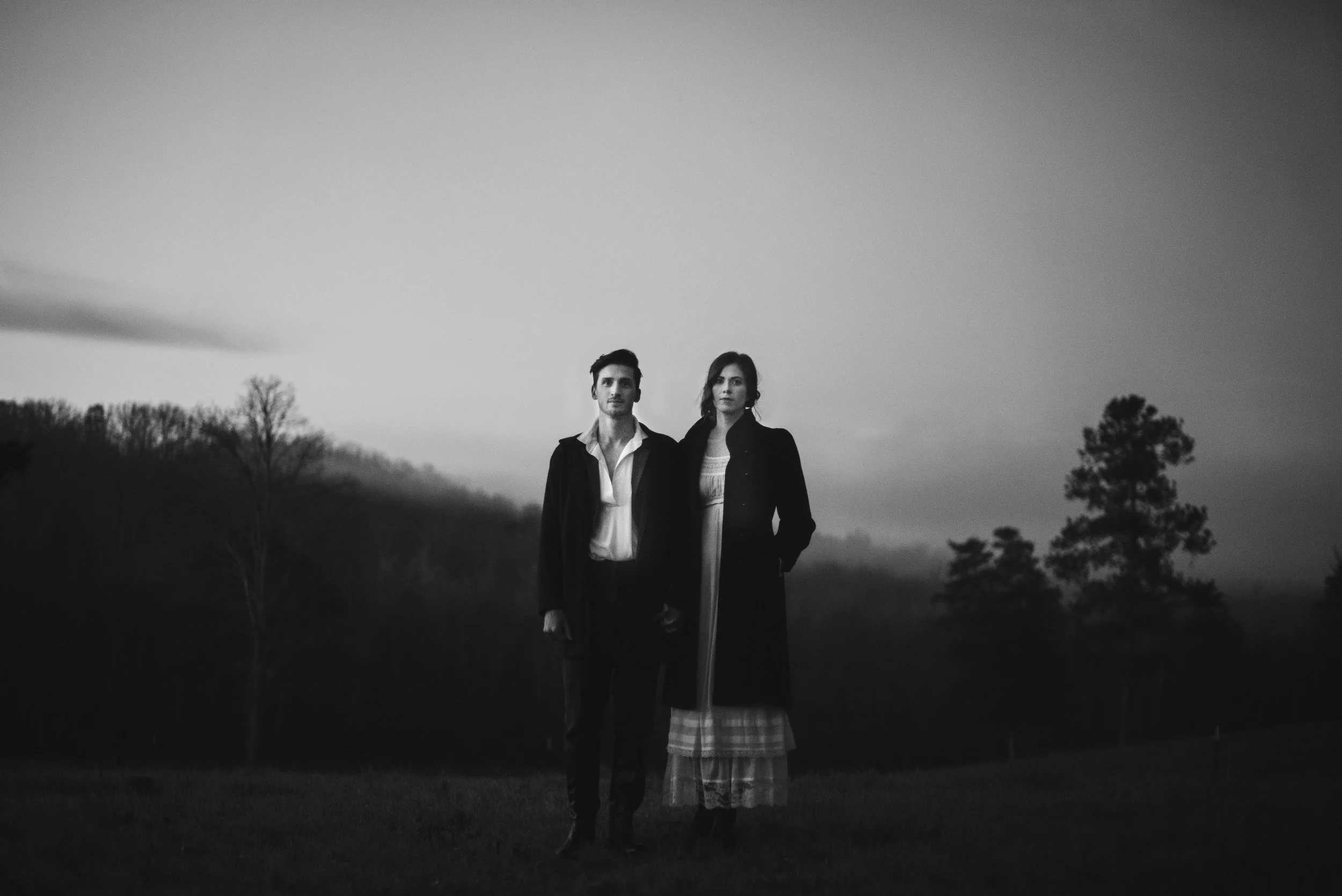 A black-and-white photo of a man and a woman standing outdoors at dusk or dawn, with trees and an open landscape in the background.