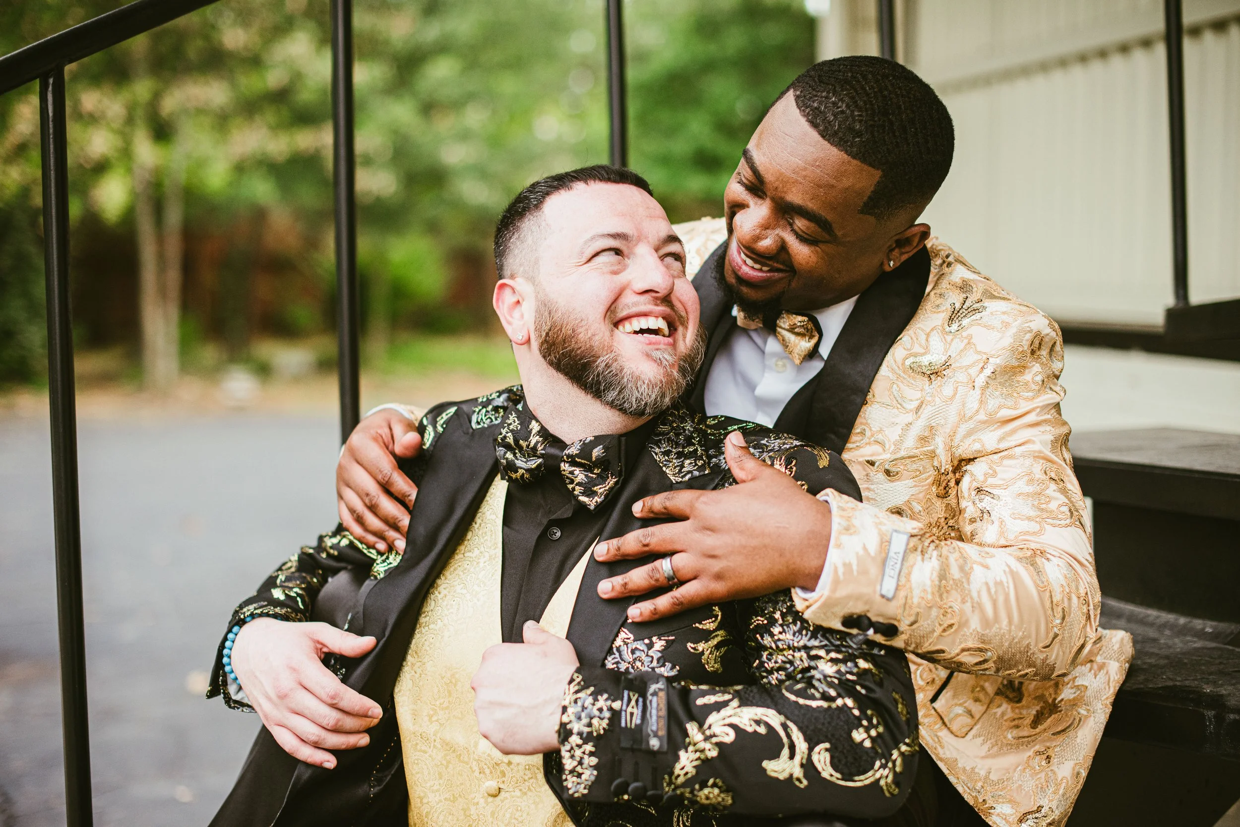 Two men dressed in ornate suits sharing a joyful hug outdoors, with one man in black and gold and the other in gold and black, surrounded by green trees.