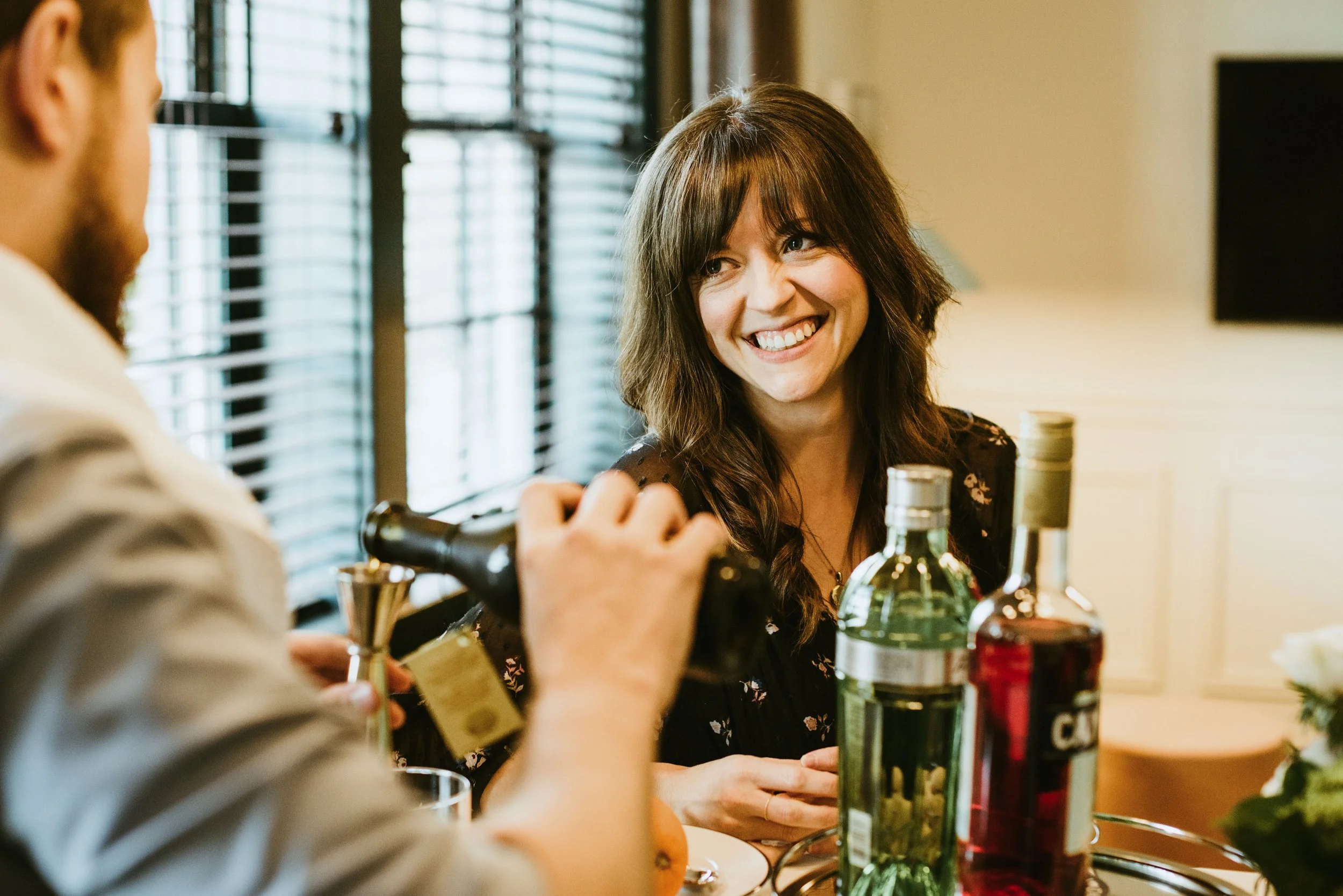 A woman with long brown hair smiling, sitting at a table with wine bottles and other items, engaged in conversation with a person blurred on the left side of the image.