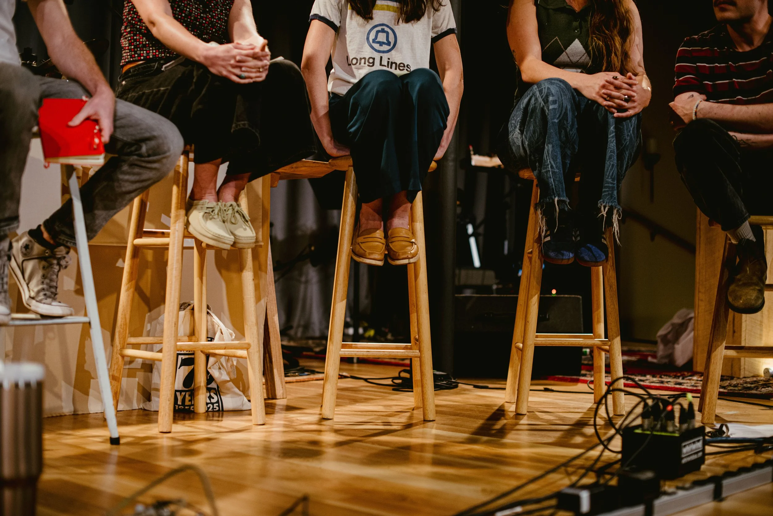 People sitting on wooden stools on a stage, with some holding items like a red notebook. The focus is on the lower half of their bodies, showing their legs, shoes, and clothing. The stage has a wooden floor, and there are some cables and equipment vi