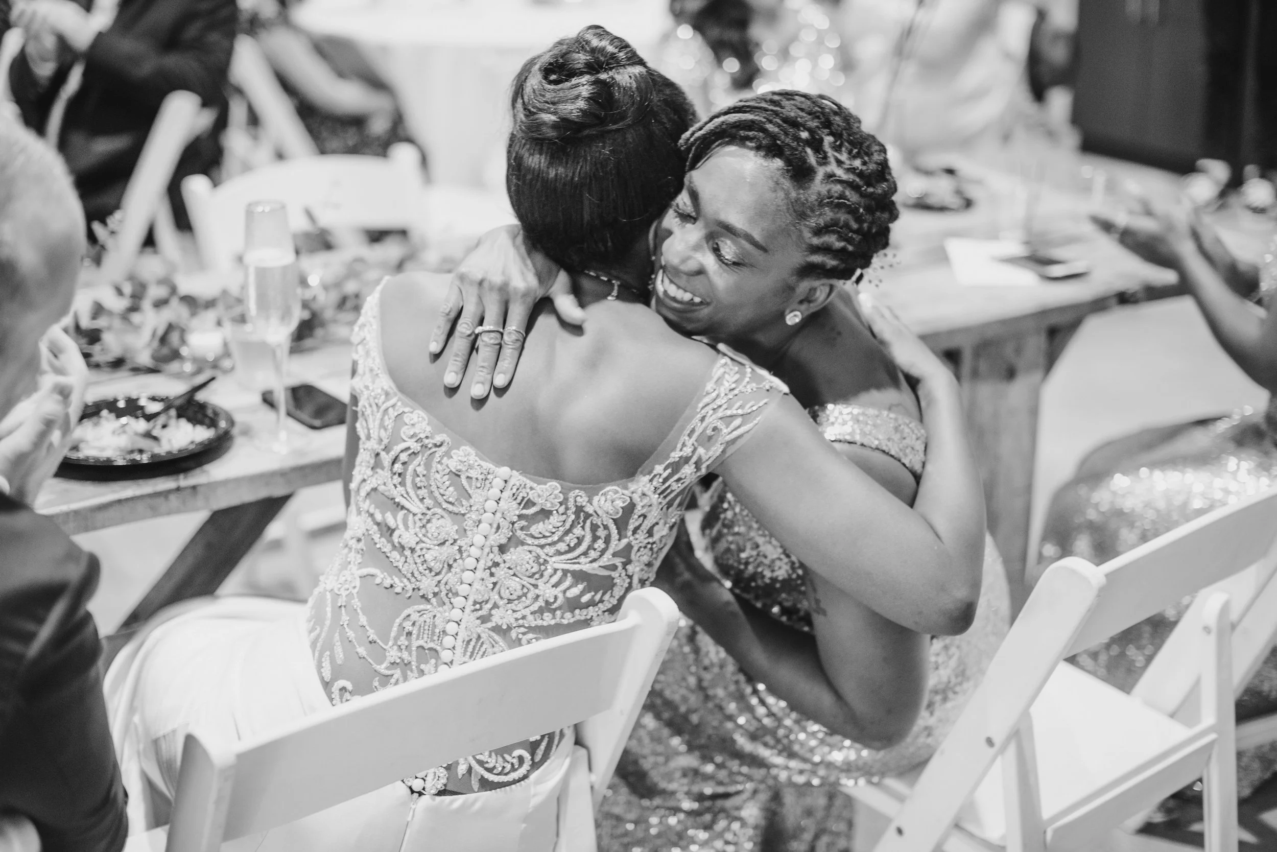Two women hugging at a celebration or gathering, smiling and embracing each other warmly, sitting at a table with food and drinks.
