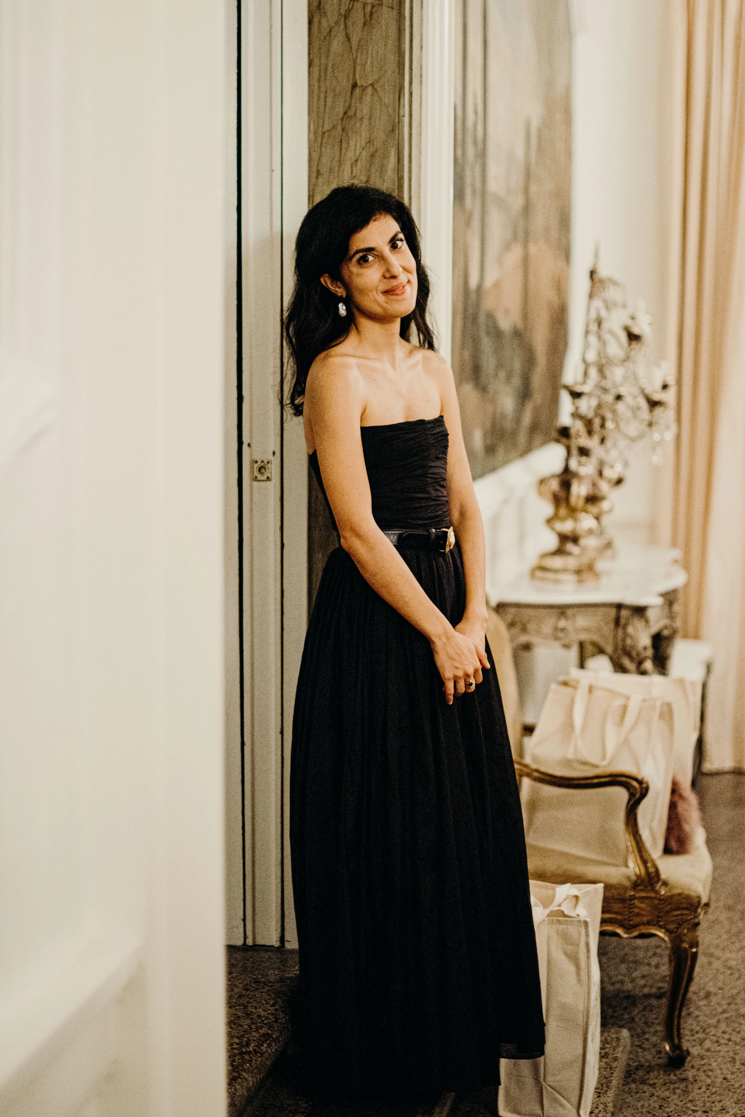 A woman in a strapless black dress standing in a room with ornate decor, including a painting, a mirror, and antique furniture.