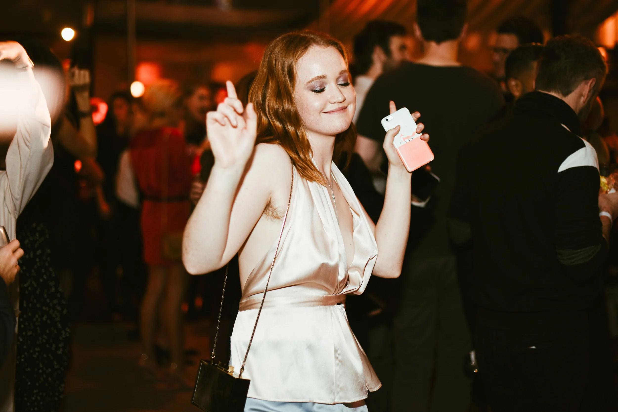 A young woman with red hair and freckles dancing at a party, holding a phone in one hand and raising the other, with her eyes closed and a smile on her face. She is wearing a sleeveless satin dress and a cross necklace, surrounded by other people in 