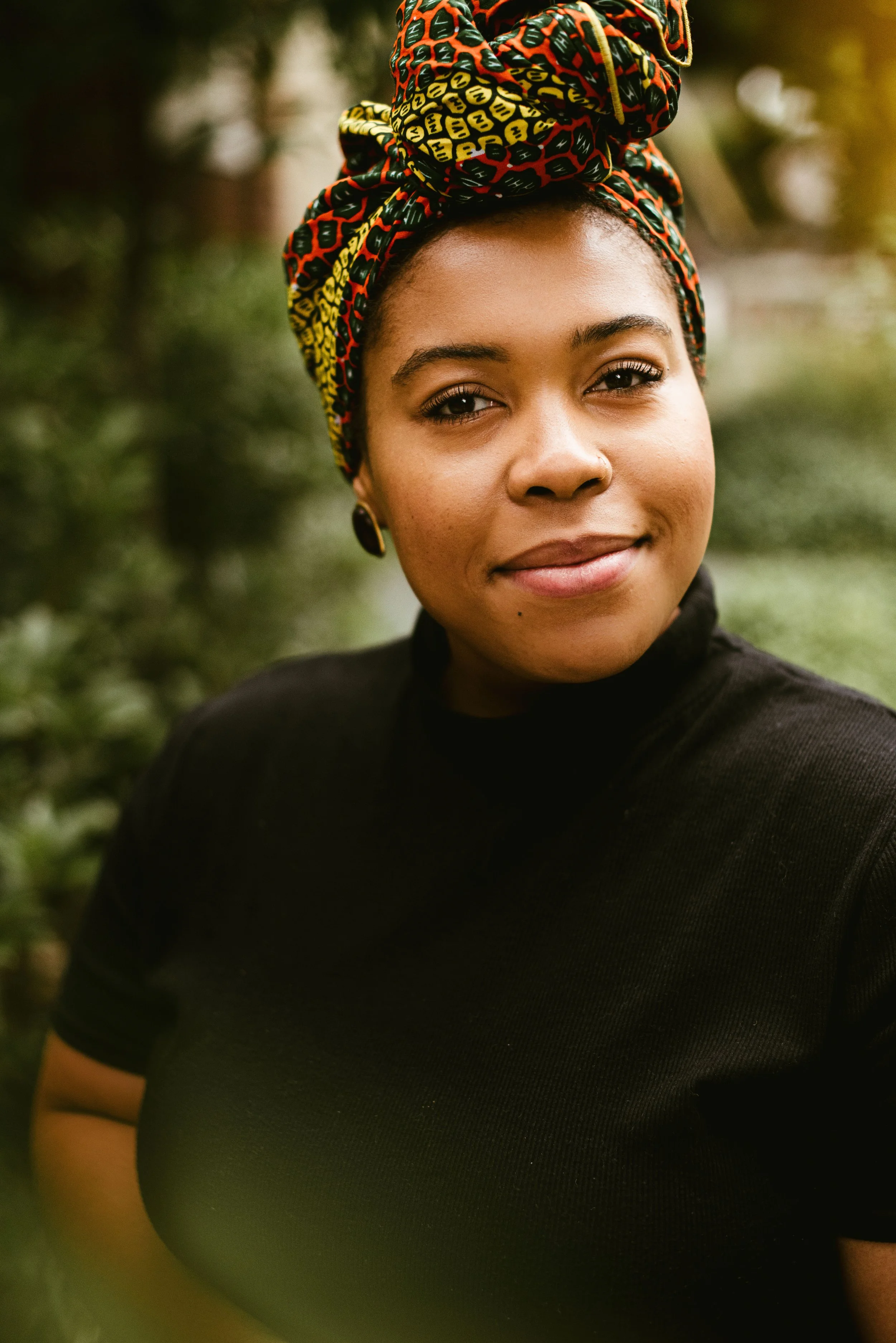 Close-up of a woman with a colorful headwrap, wearing a black top, outdoors with blurred nature background.