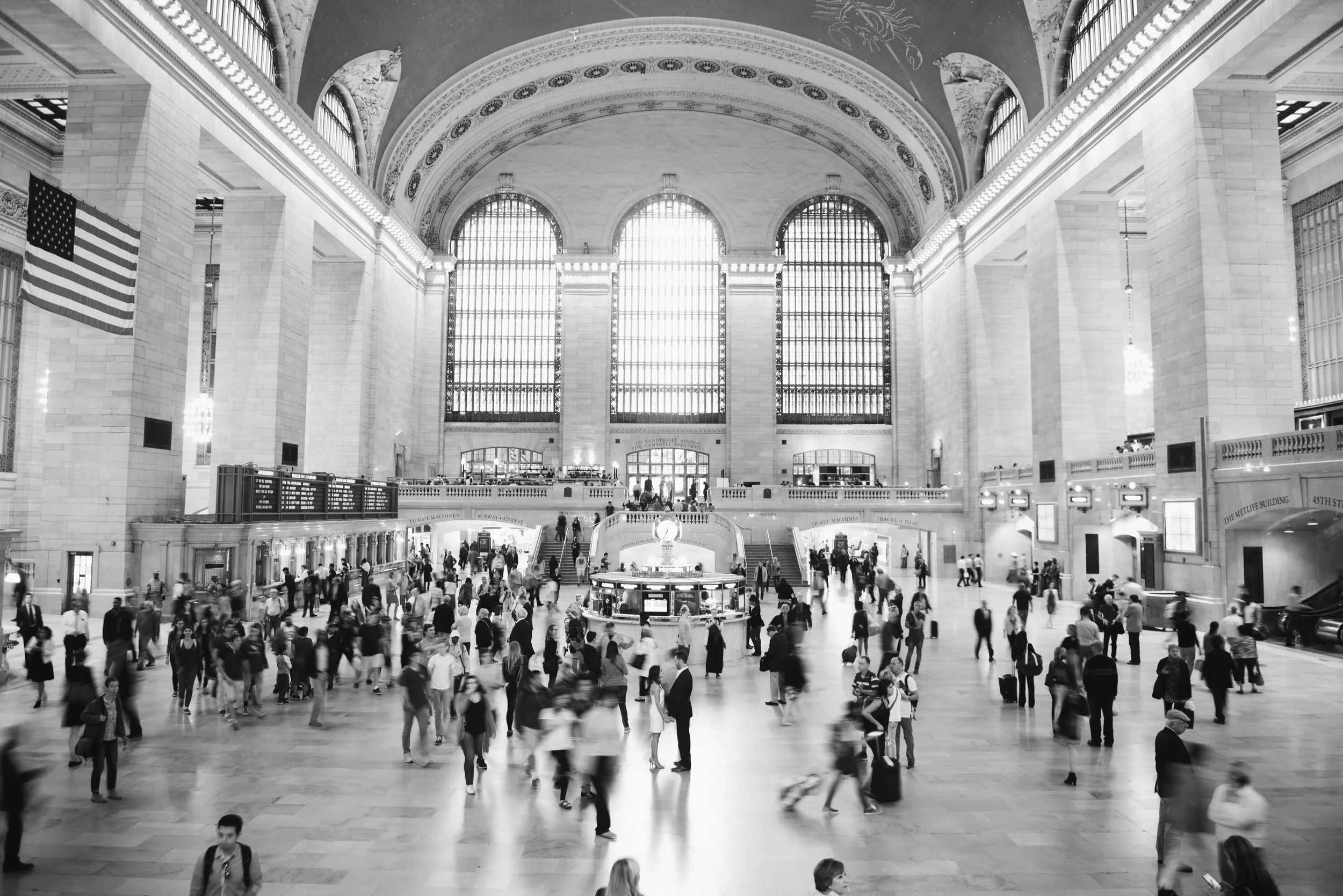 The main hall of a historic train station with high arched windows, large American flag hanging on the wall, and many people walking and standing around.