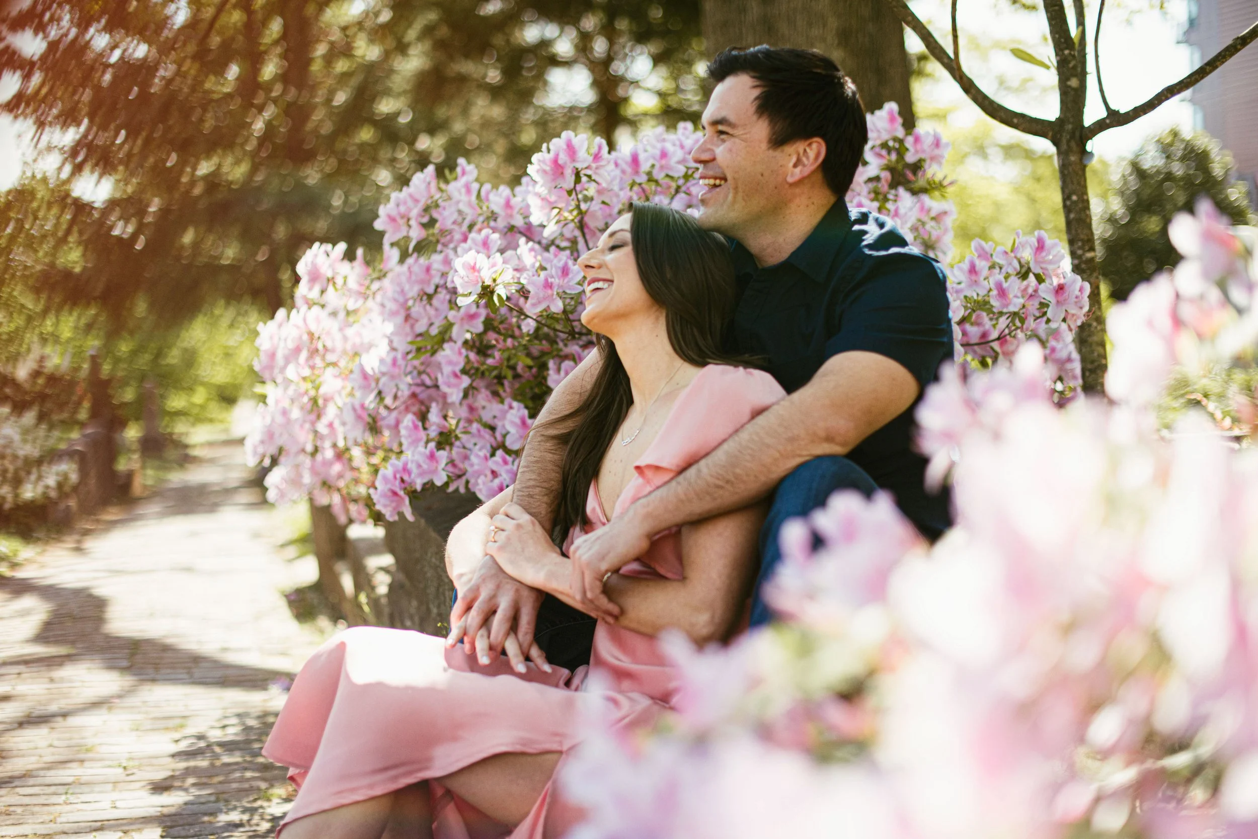 A man and woman sitting on a park bench surrounded by pink flowers, smiling and enjoying each other's company.