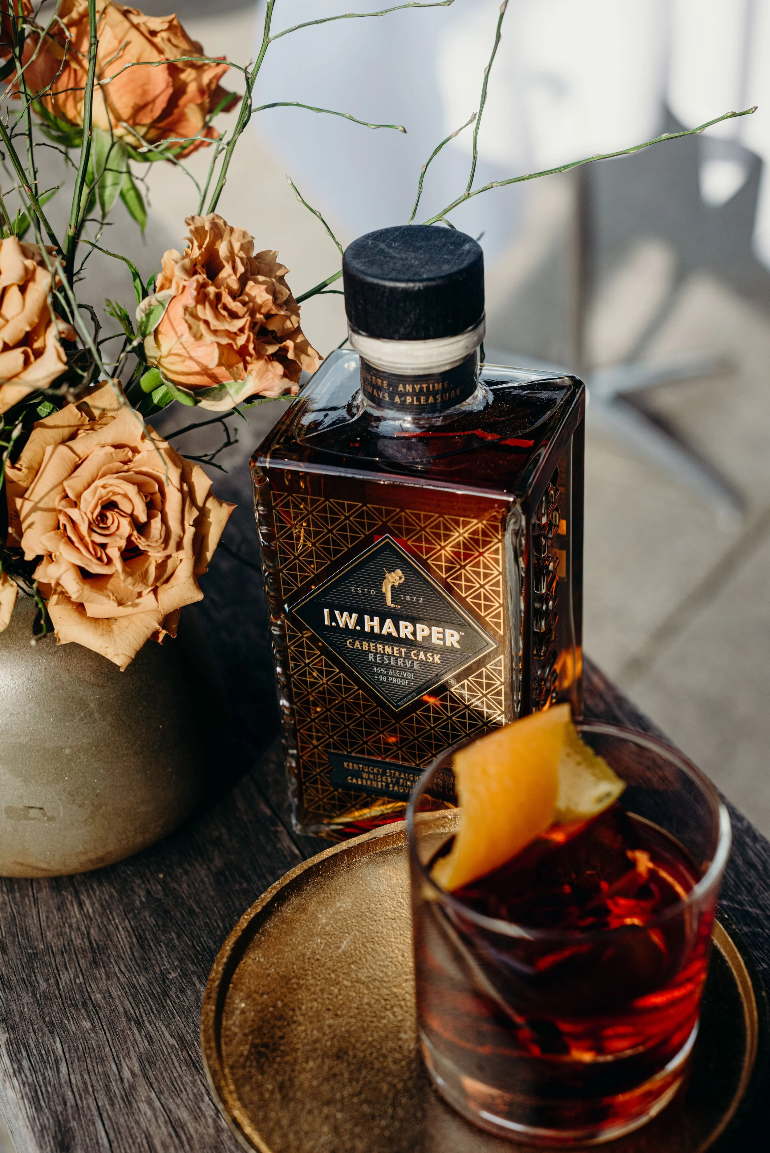 A bottle of I.W. Harper bourbon whiskey next to a glass of whiskey with a lemon twist, placed on a dark wooden table, with dried roses and a gold-rimmed plate nearby.