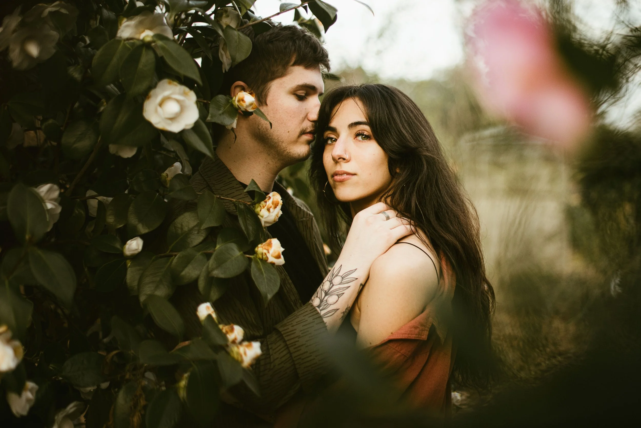 A young man and woman hugging among green foliage and white flowers, with their faces close together and the woman looking at the camera.