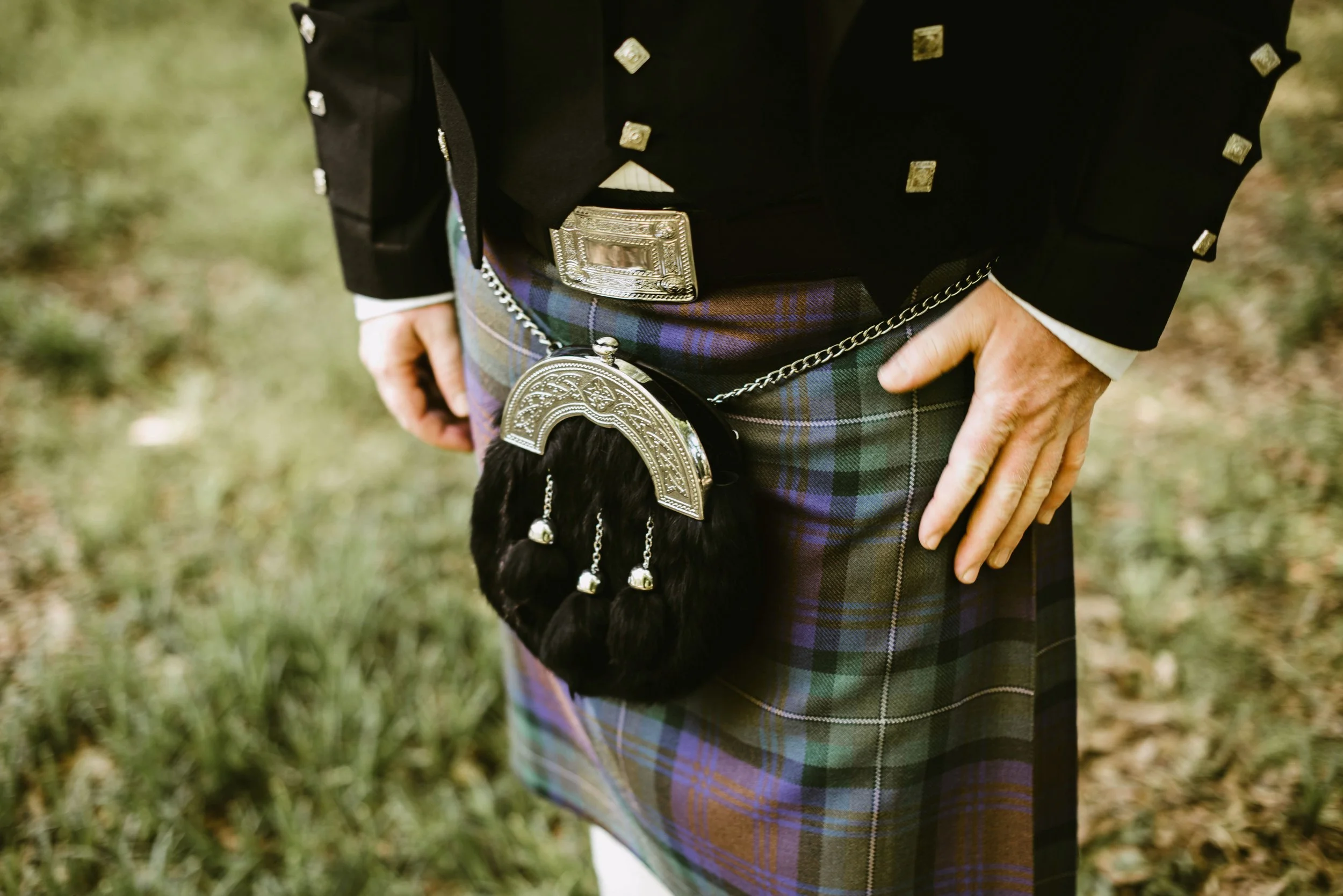 A person wearing traditional Scottish attire, including a kilt with a plaid pattern, a black jacket with silver embellishments, a wide decorated belt, and a sporran with a chain.