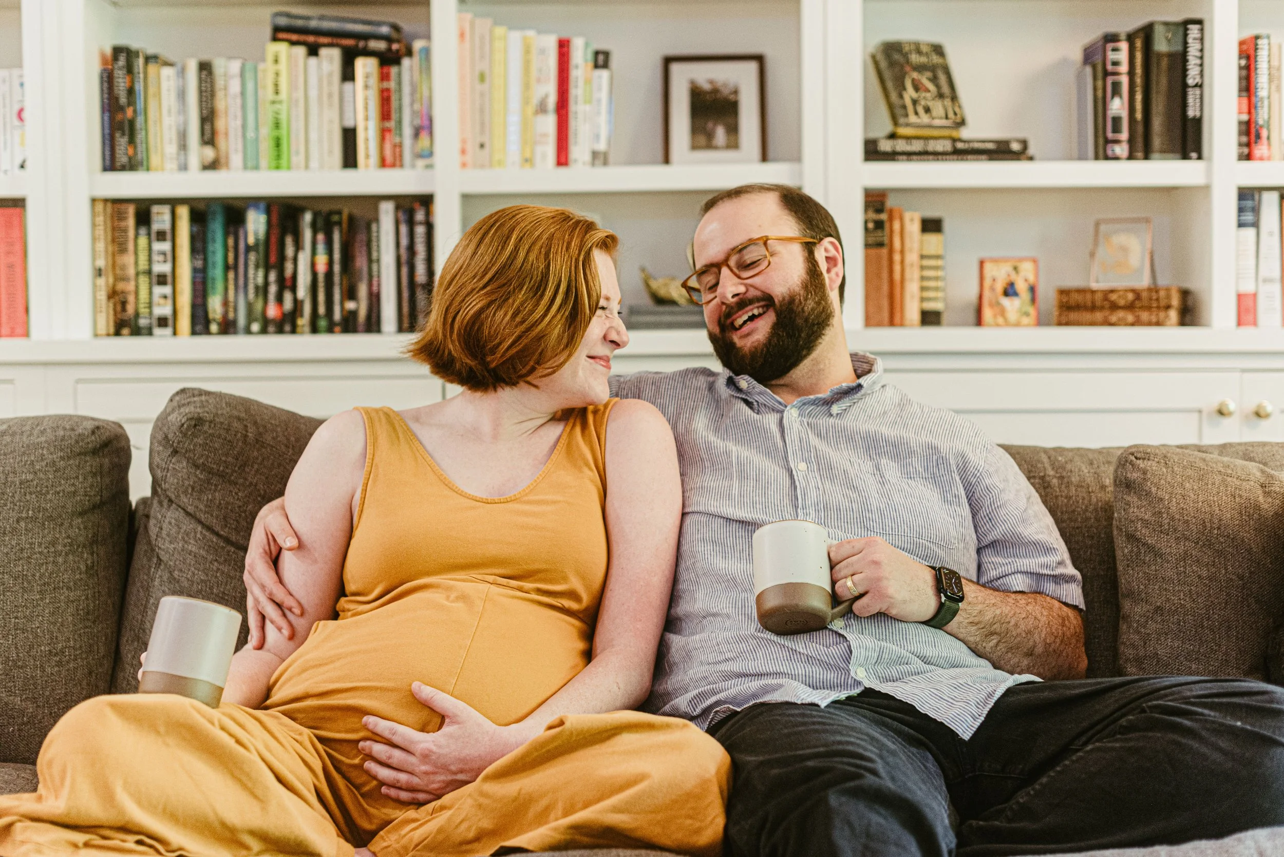 A couple sitting on a couch sharing a laugh, the woman has red hair and is wearing an orange dress, the man has a beard, glasses, and is wearing a striped shirt holding a coffee mug. They are in a cozy room with a bookshelf in the background.
