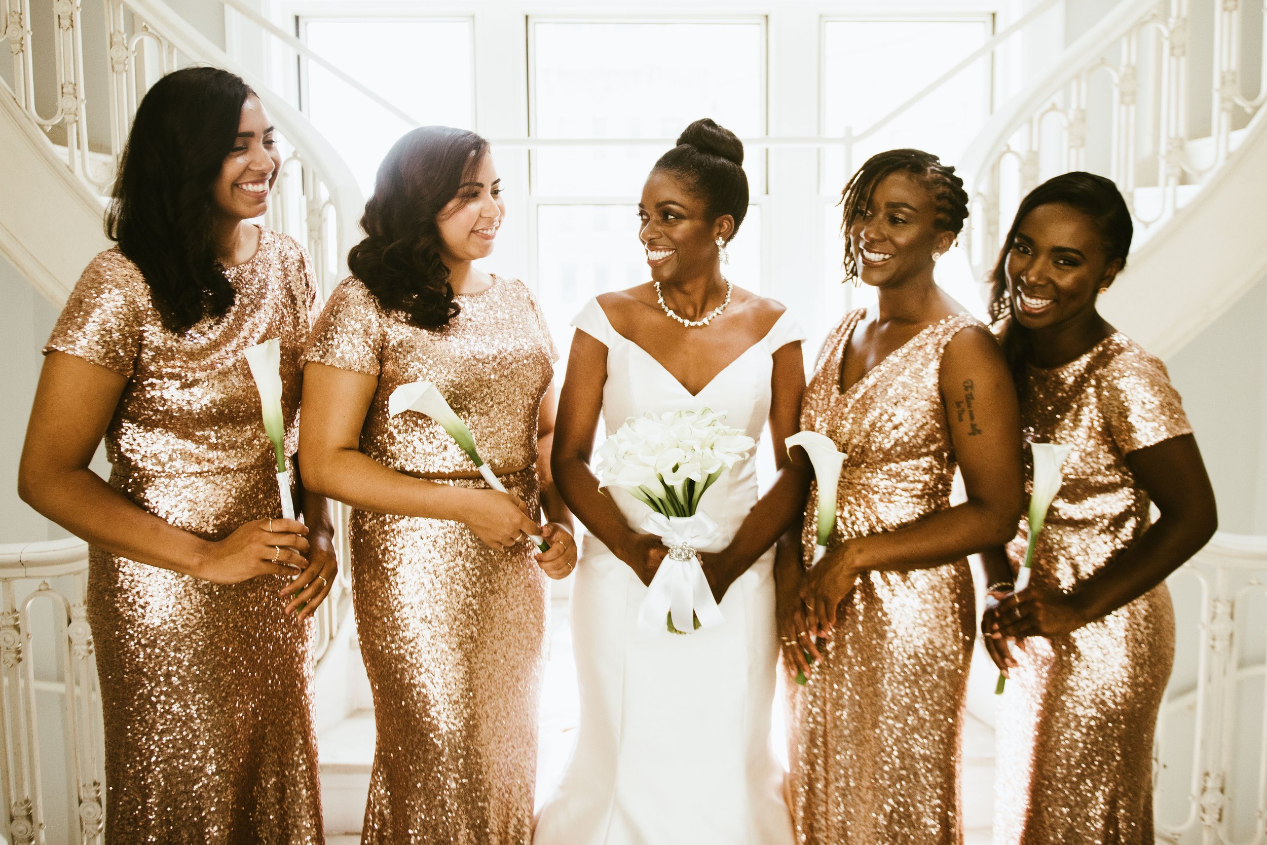 A bride and five bridesmaids in sequined dresses standing together indoors near a staircase, smiling and holding white calla lily bouquets.