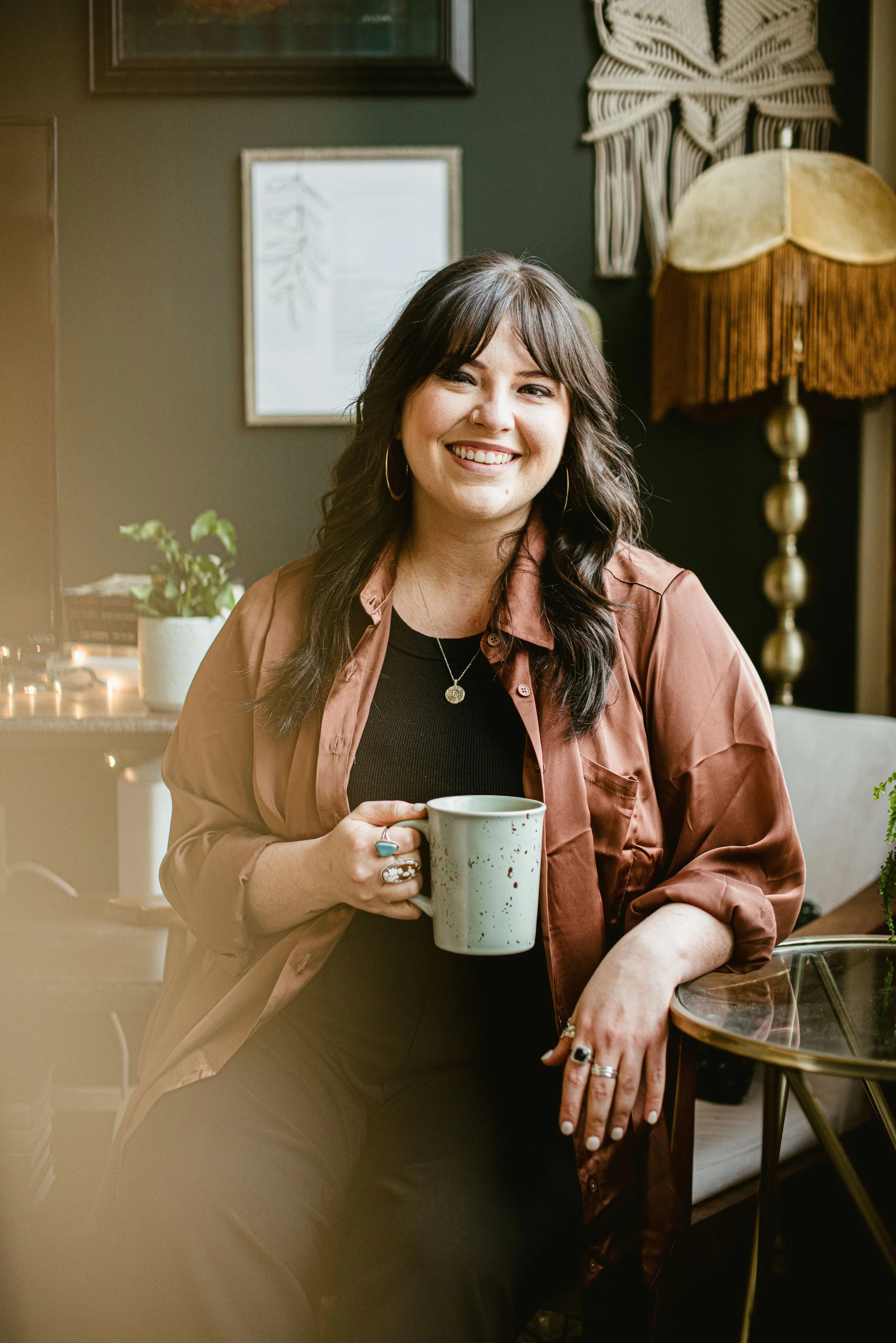 A smiling woman with dark hair holding a white mug, sitting in a cozy, well-decorated room with a dark wall, a lamp, and a plant in the background.