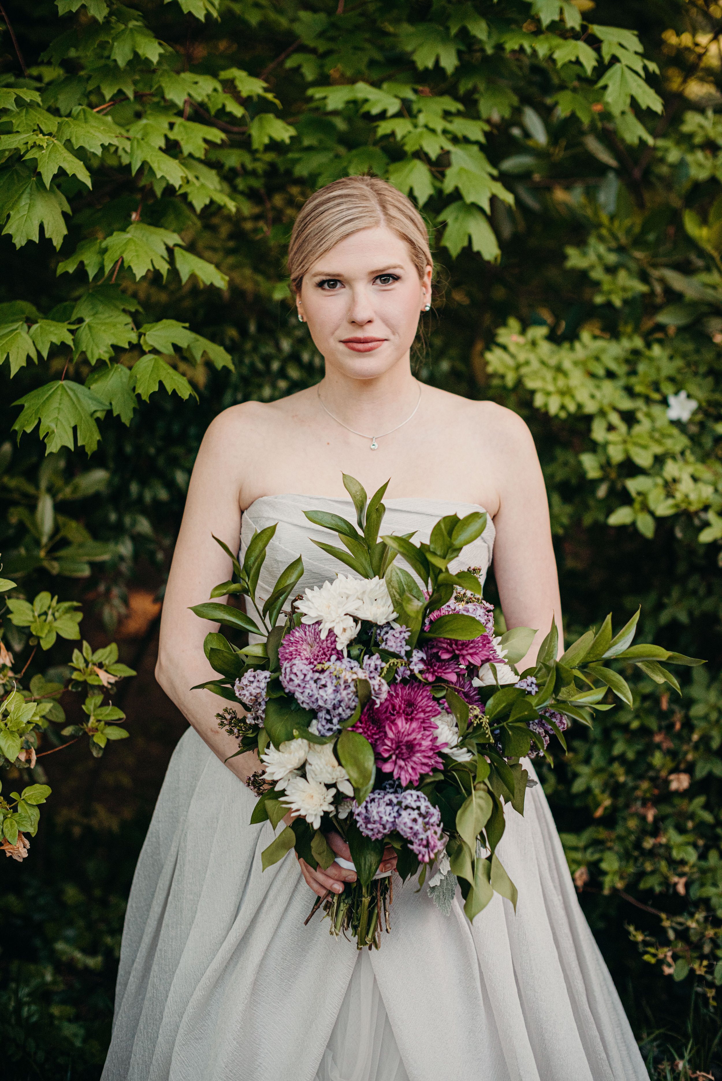 A woman in a strapless wedding dress holding a large bouquet of white, purple, and pink flowers with green leaves, standing outdoors in front of a background of green leaves.