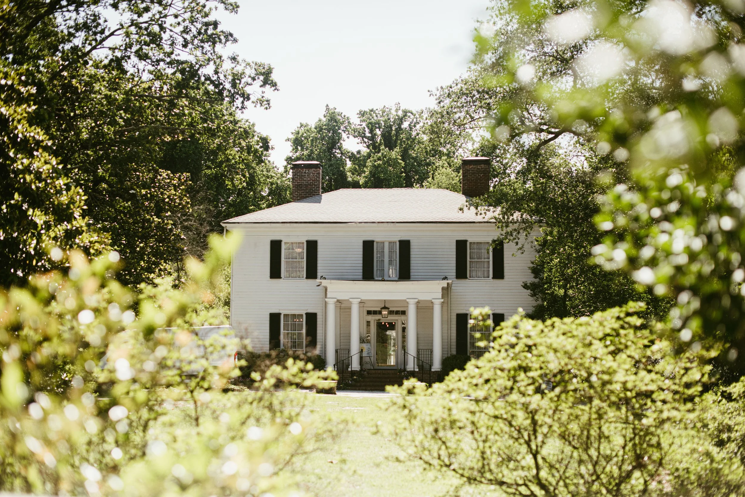 A white two-story house with black shutters, visible through green bushes and trees, with a brick chimney and a porch with white columns.