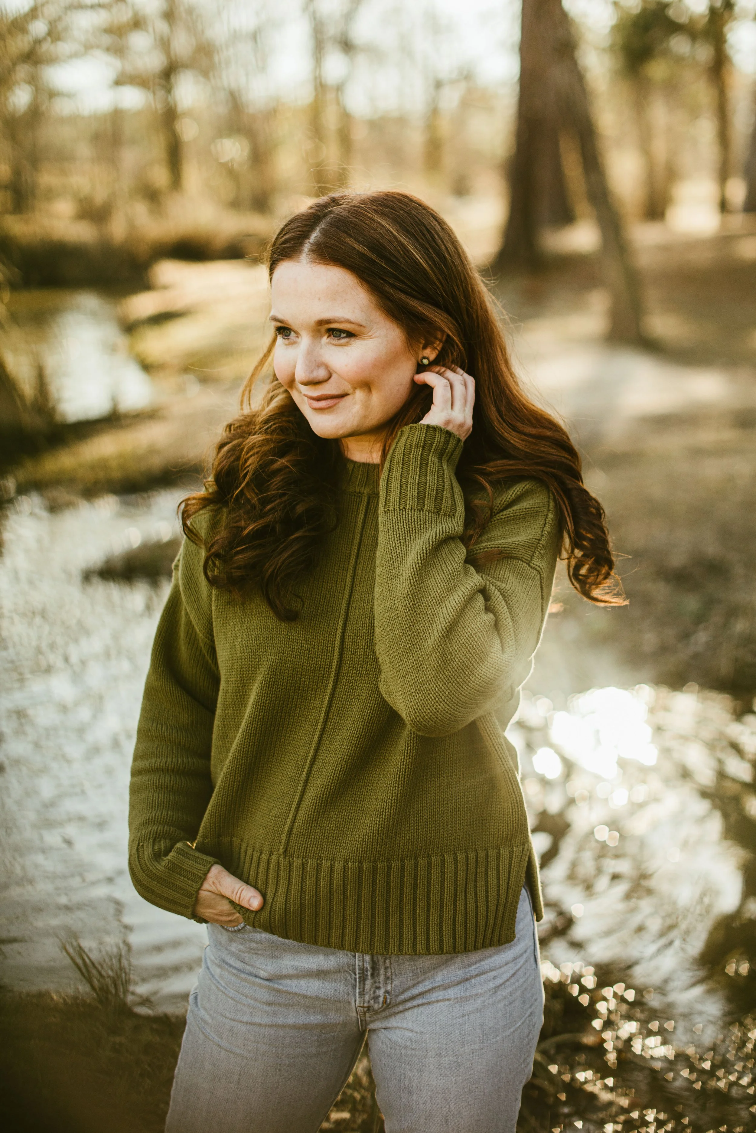 A woman with long, wavy reddish-brown hair standing outdoors by a river or creek with trees in the background, wearing a green zip-up sweater and gray jeans, smiling and touching her ear.