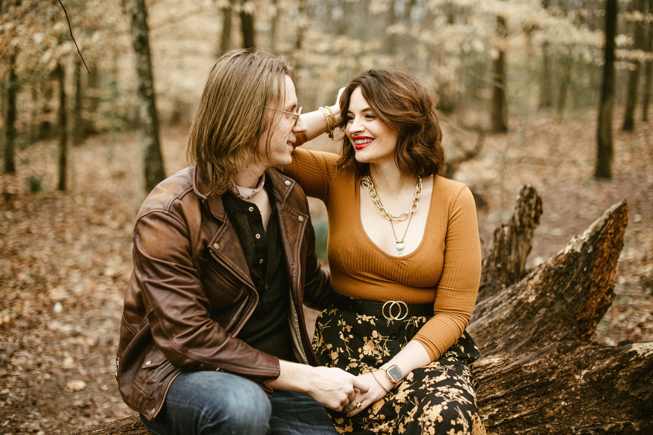 A couple sitting on a fallen log in a forest, smiling and gazing at each other, with autumn leaves around them.