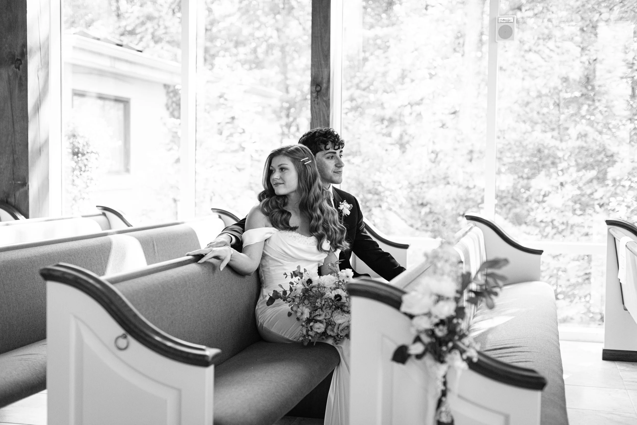 A bride and groom sitting on pews inside a church during a wedding ceremony, with the bride holding a bouquet of flowers and the groom sitting closely beside her.