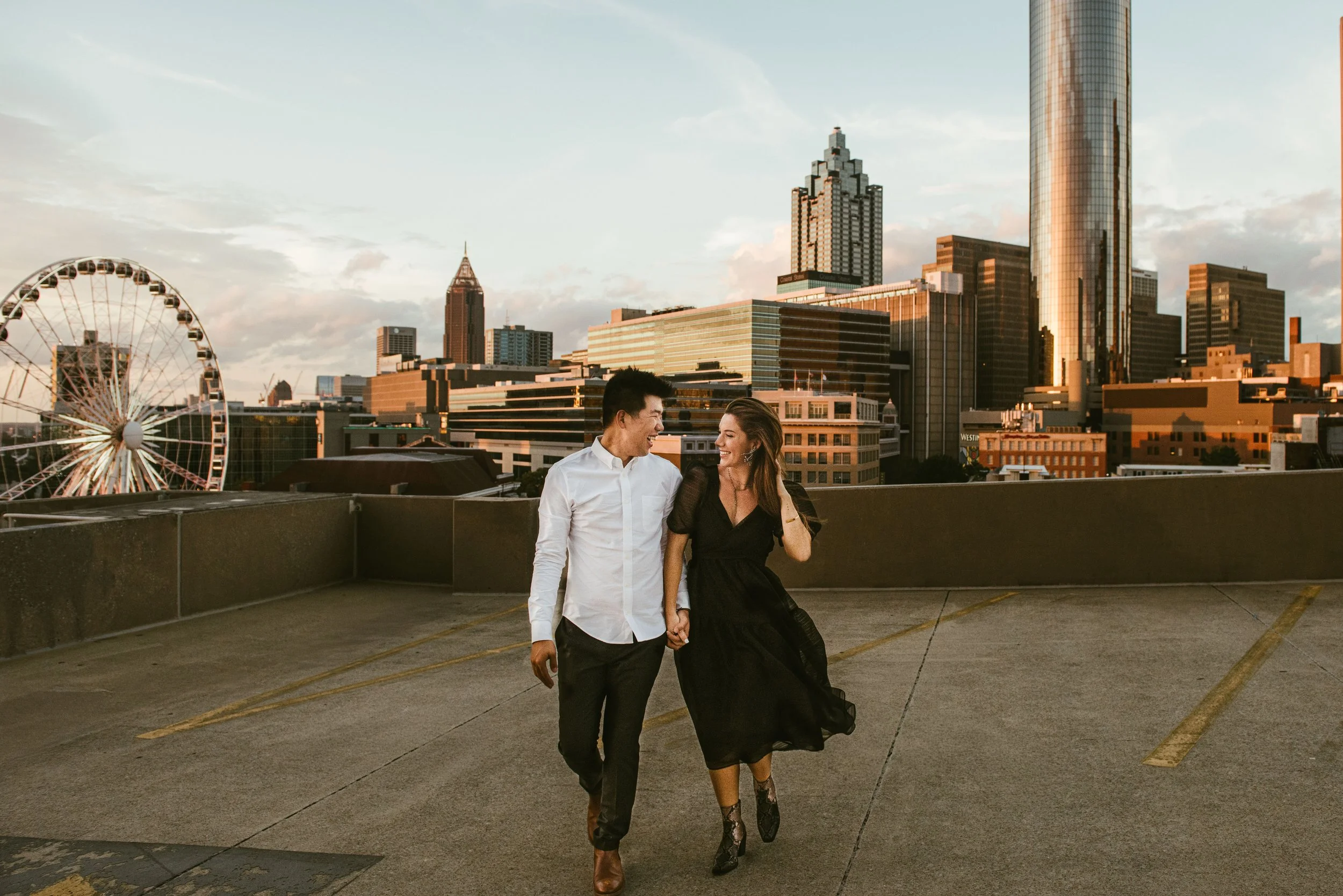 A man and woman walking hand in hand in a parking lot with a city skyline in the background during sunset.
