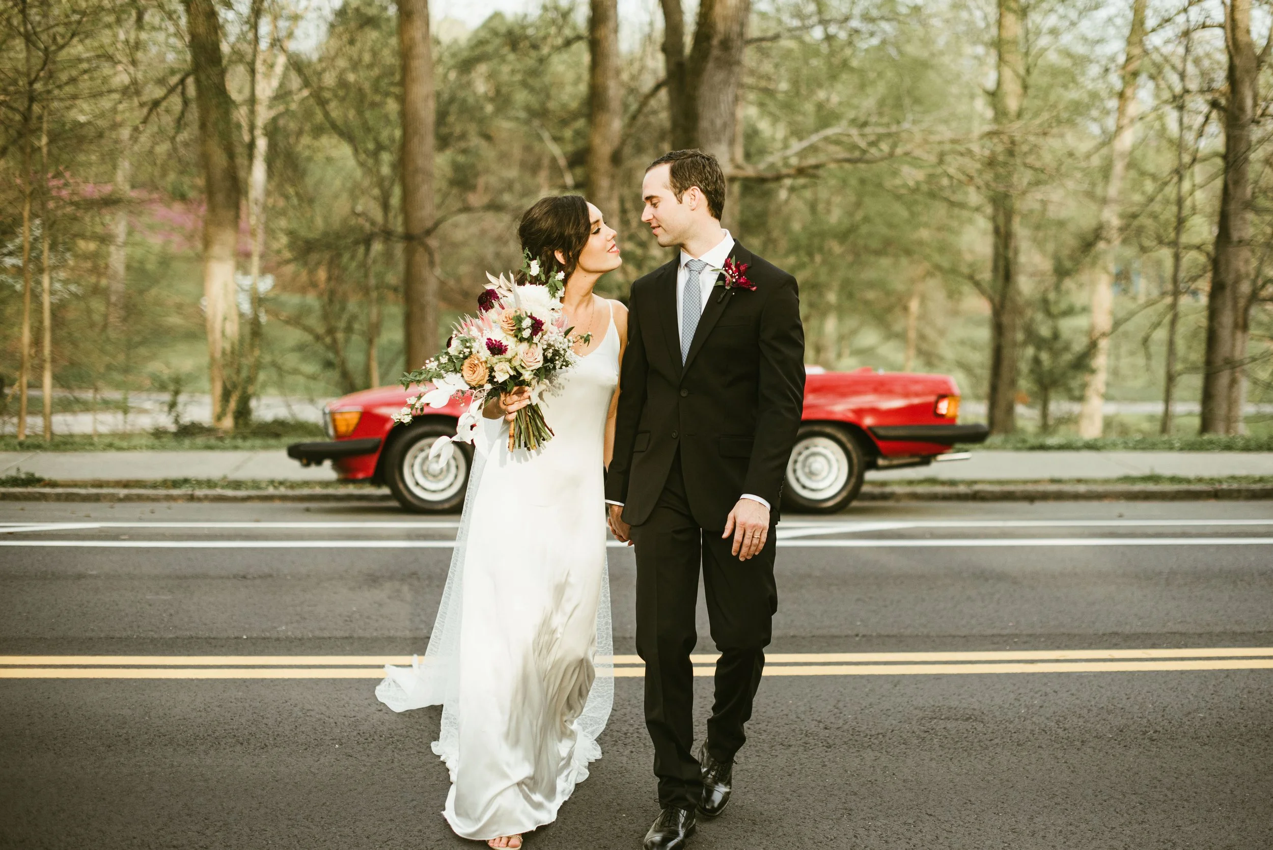 A bride and groom holding hands on a street, surrounded by trees, with a red vintage car parked behind them.