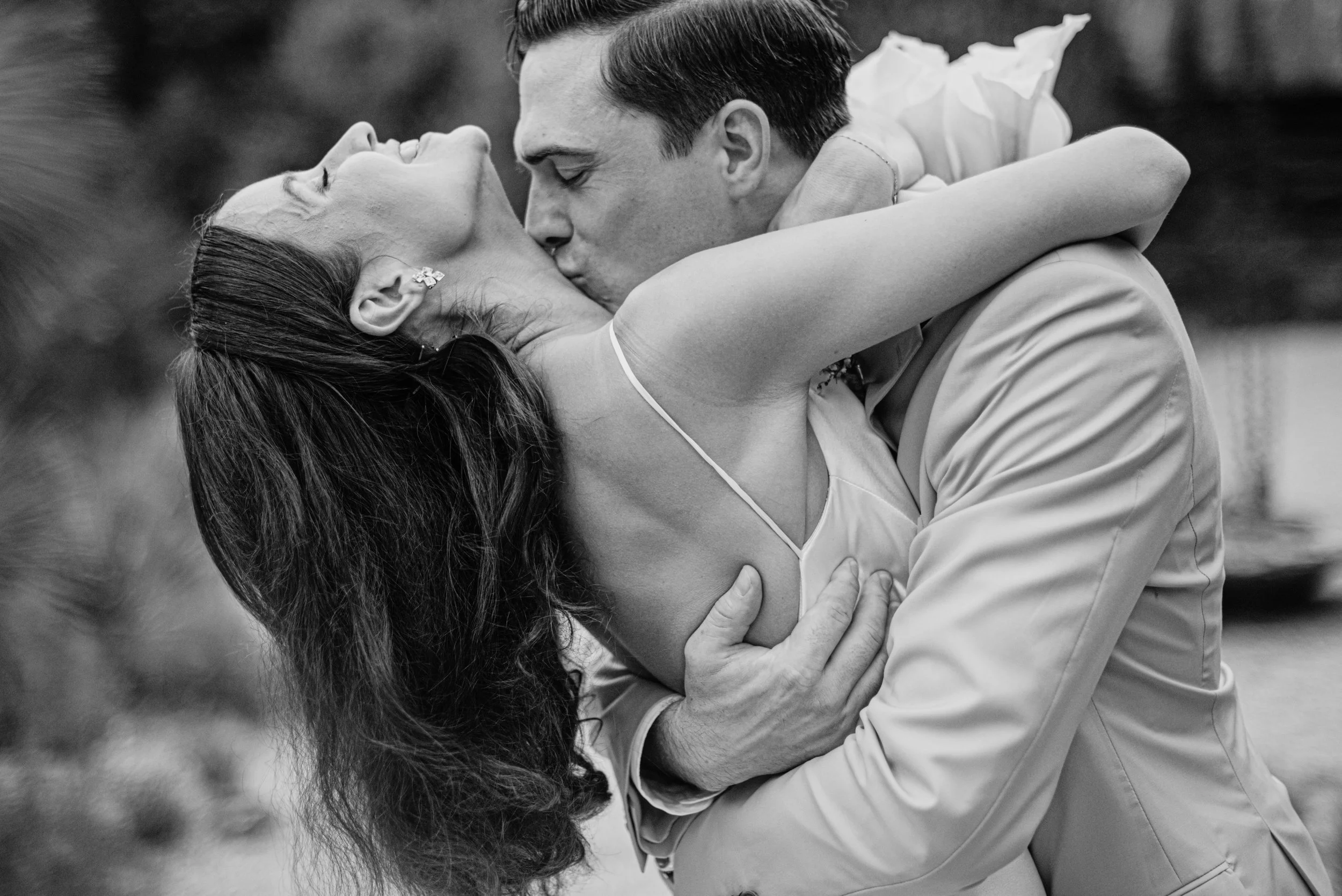 A black-and-white photograph of a couple sharing a kiss, with the woman dressed in a wedding gown and the man in formal attire. The woman has long hair and is wearing earrings, while the man has short hair. They are embracing each other tenderly.