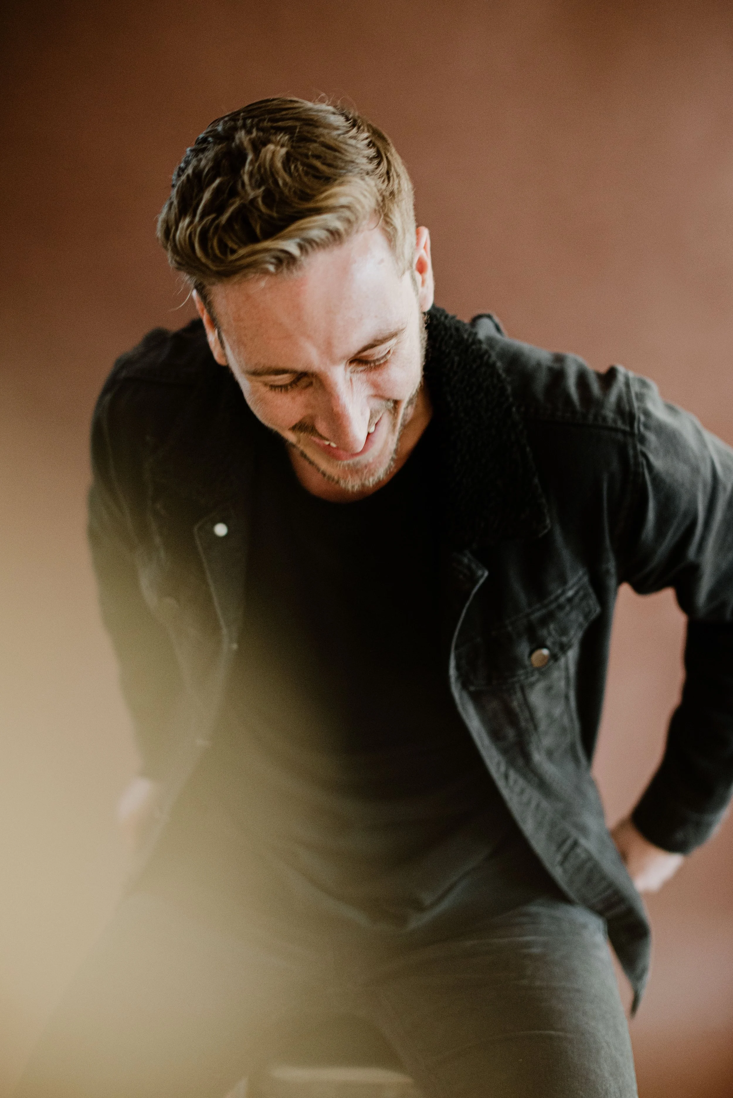 A man with light brown hair styled to the side, wearing a black shirt and a black jacket with a fuzzy collar, smiling and looking down, against a plain light brown background.