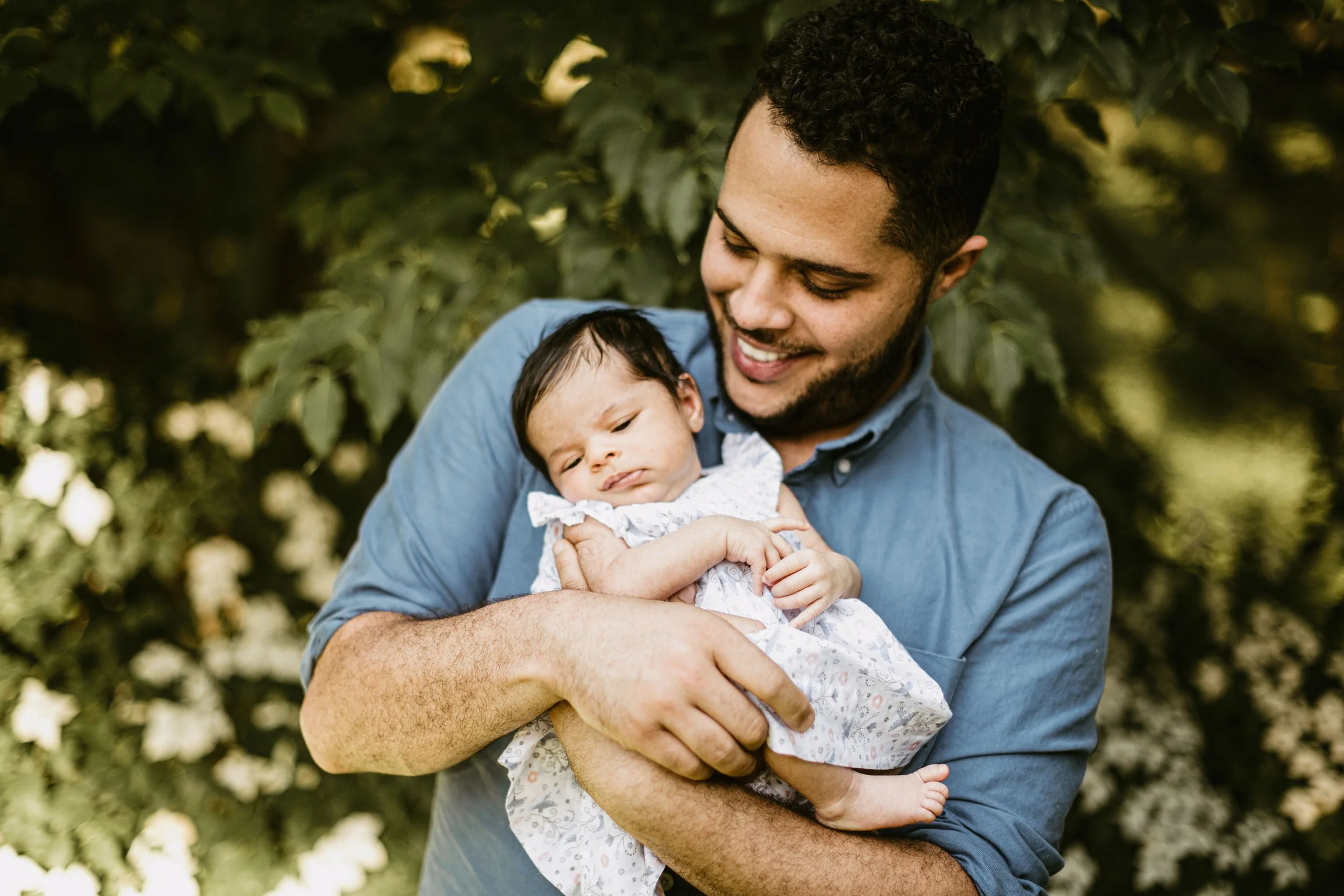 A man holding a baby outdoors with green foliage in the background.