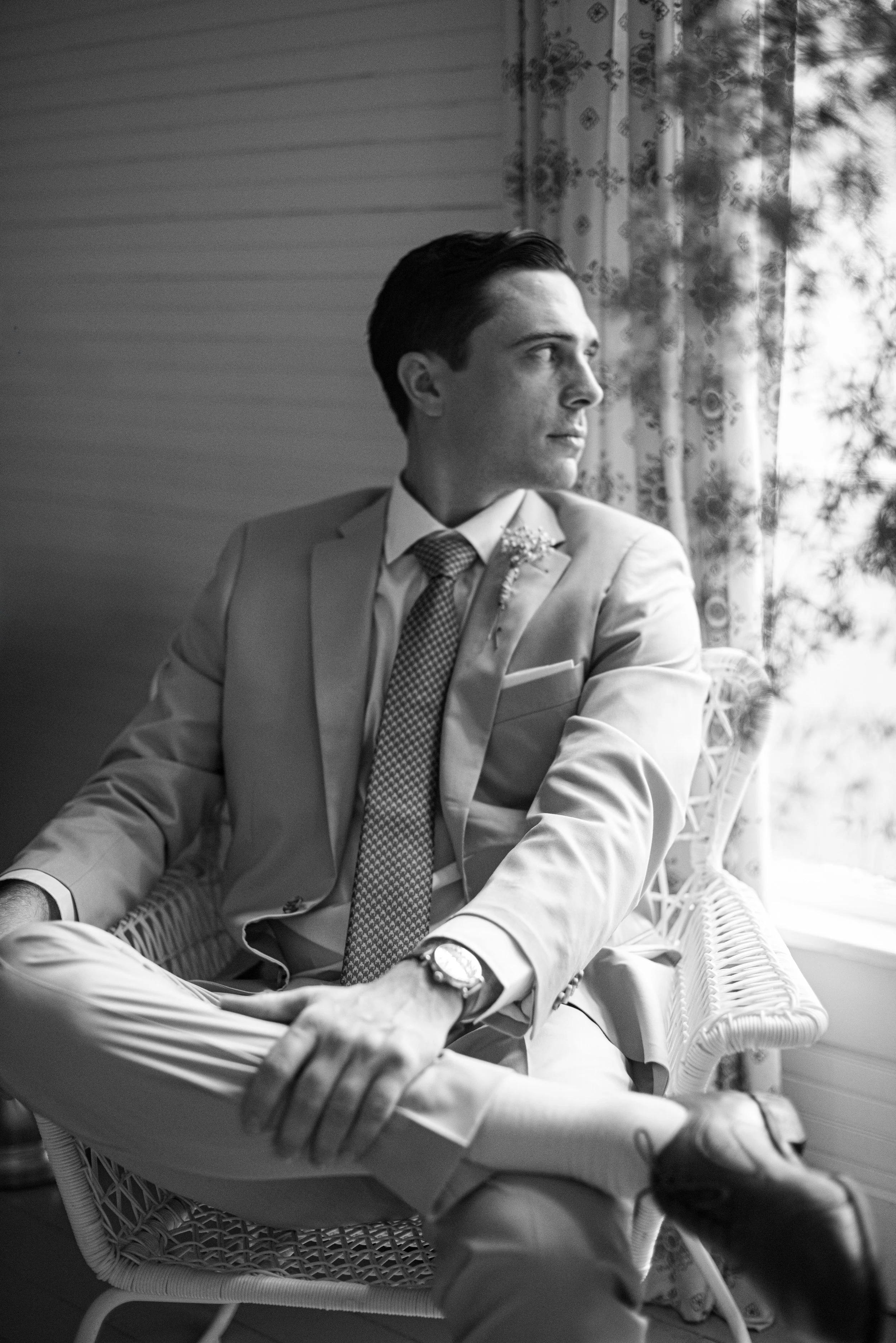A young man dressed in a suit with a tie, sitting on a wicker chair near a window with floral curtains, looking to the side.