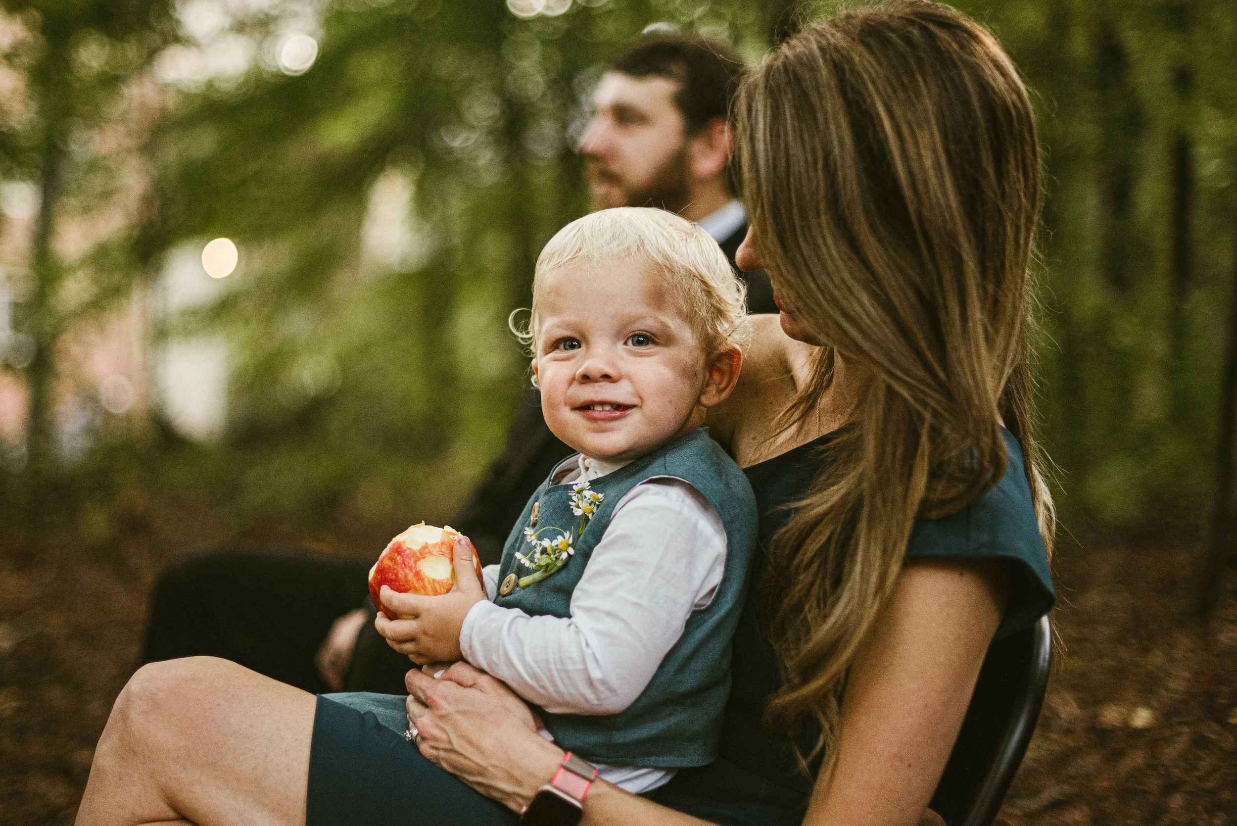 A woman with long brown hair holding a young blonde-haired child with blue eyes, sitting outdoors in a forested area. The woman is in profile, and the child is smiling while holding a red and yellow apple.